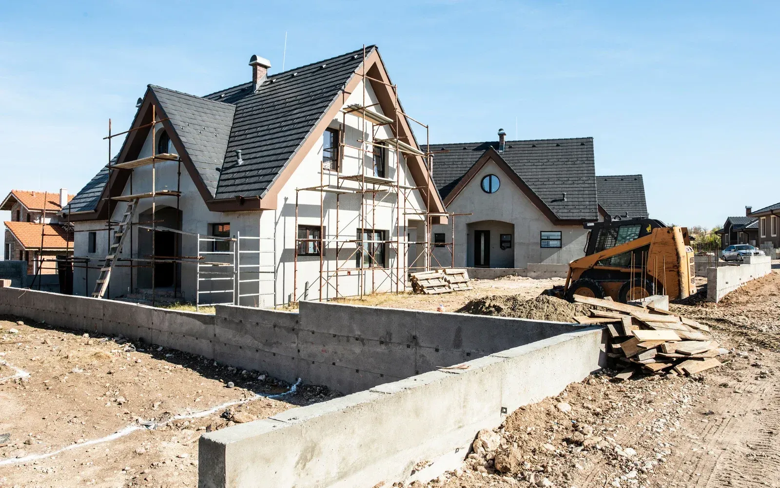 Houses under construction, with scaffolding, concrete walls, and a small excavator on a dusty lot.