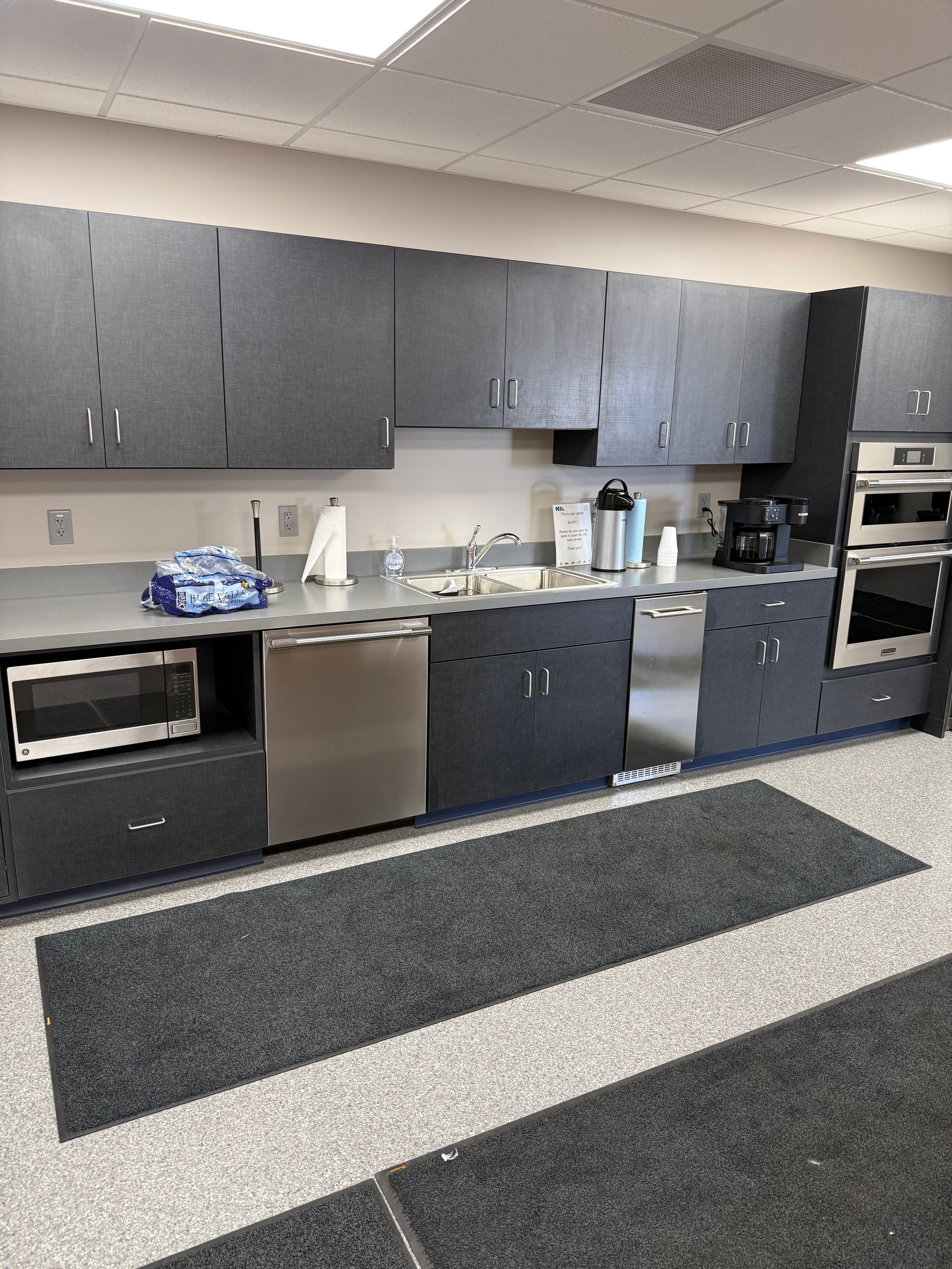 Office kitchenette with dark cabinets, microwave, sink, dishwasher, and countertop appliances.