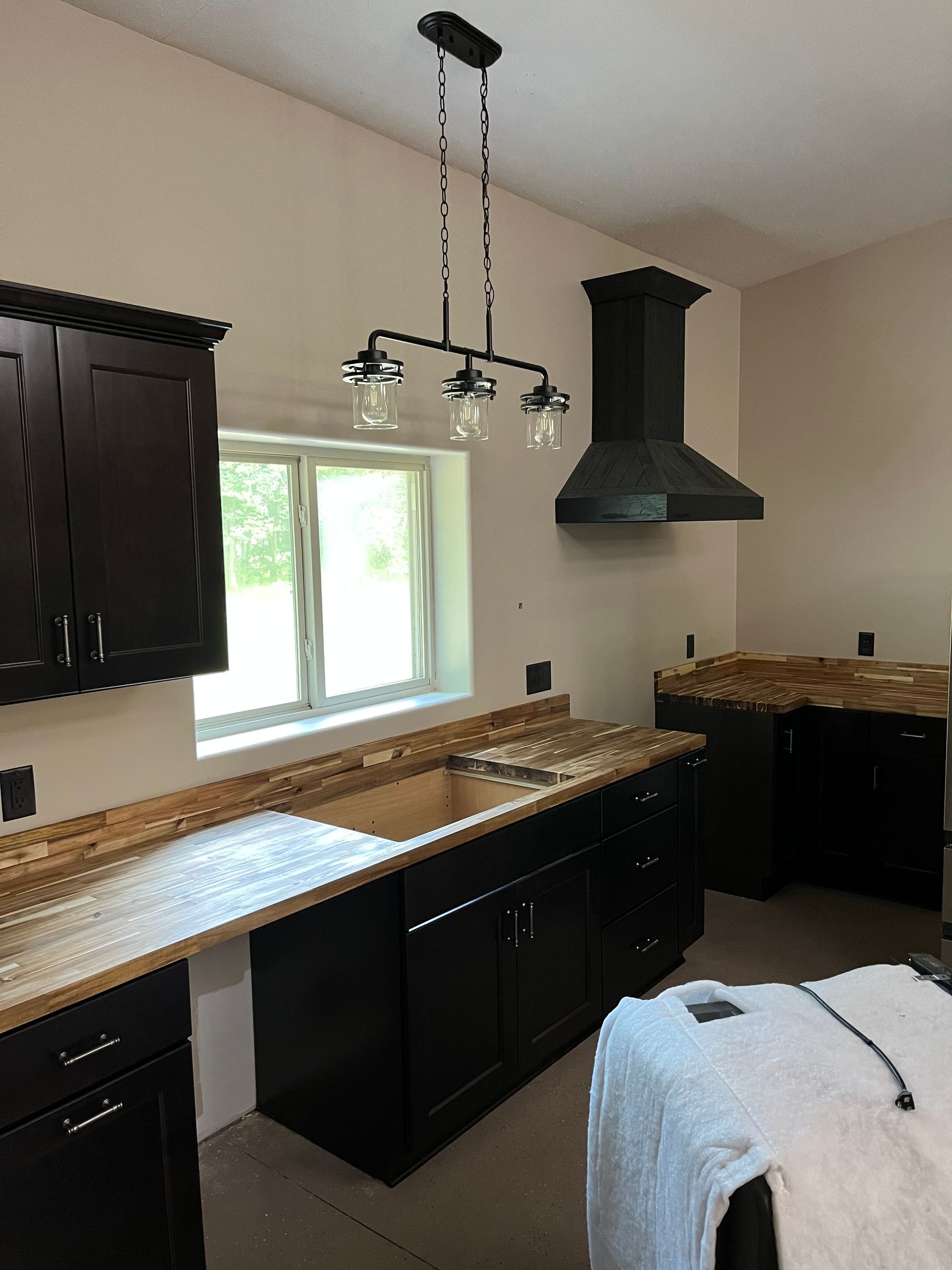 Kitchen with dark cabinets, wood countertops, a window, hanging light fixture, and black range hood.