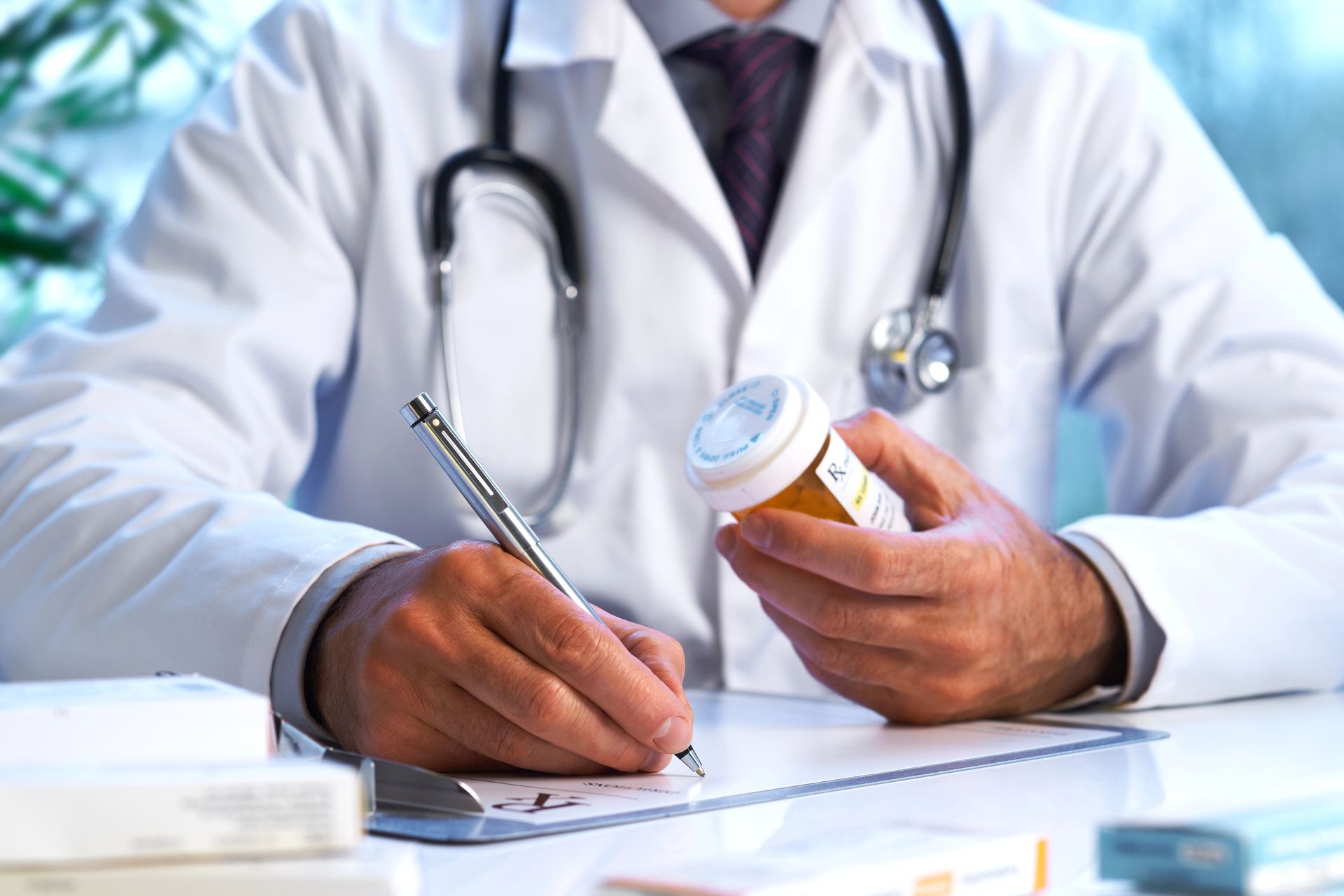 Medico in camice bianco che scrive la ricetta, tenendo in mano un flacone di pillole. Stetoscopio al collo.
