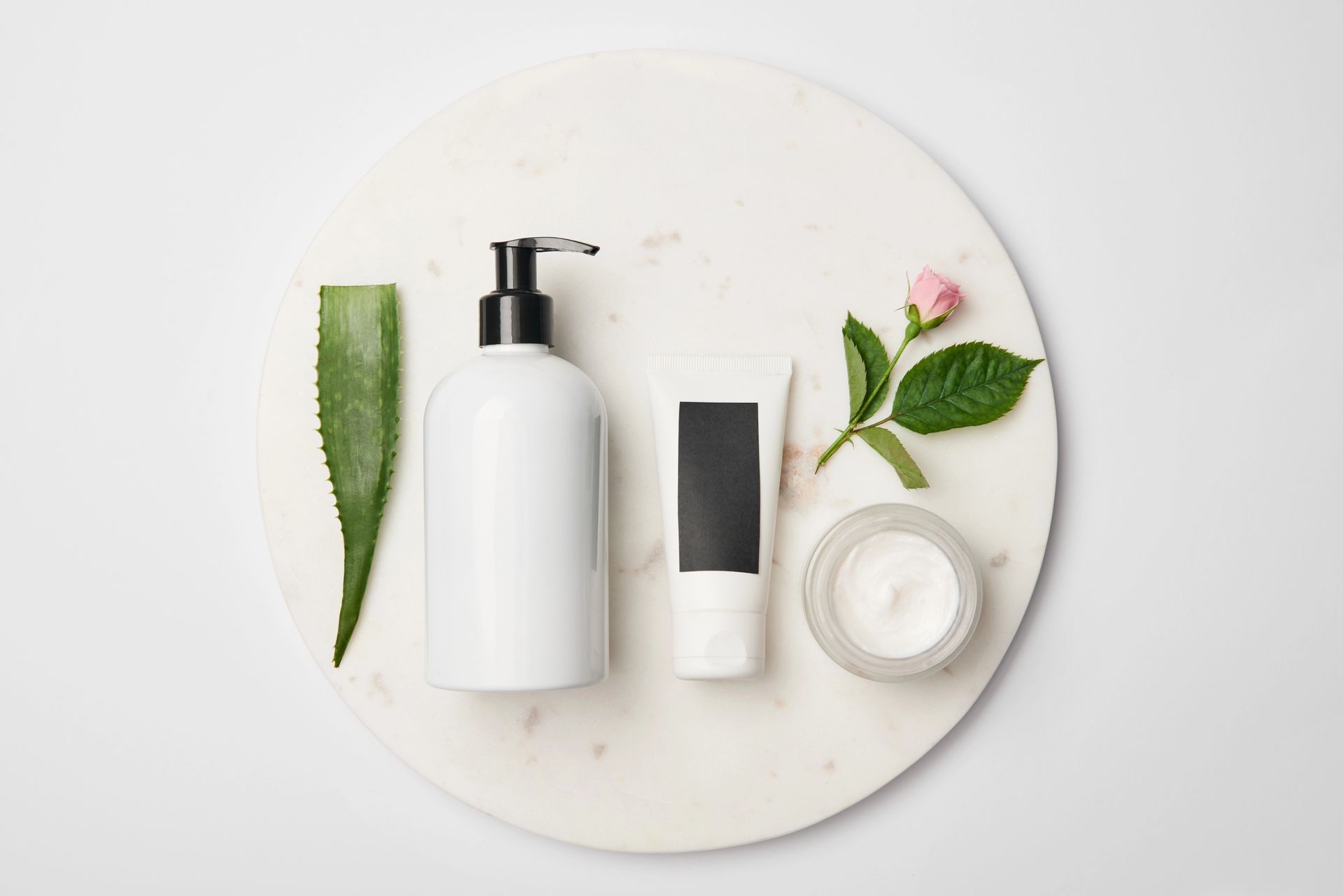 Cosmetici disposti su una superficie di marmo: flacone con pompa, tubetto, barattolo di crema, foglia di aloe e rosa.