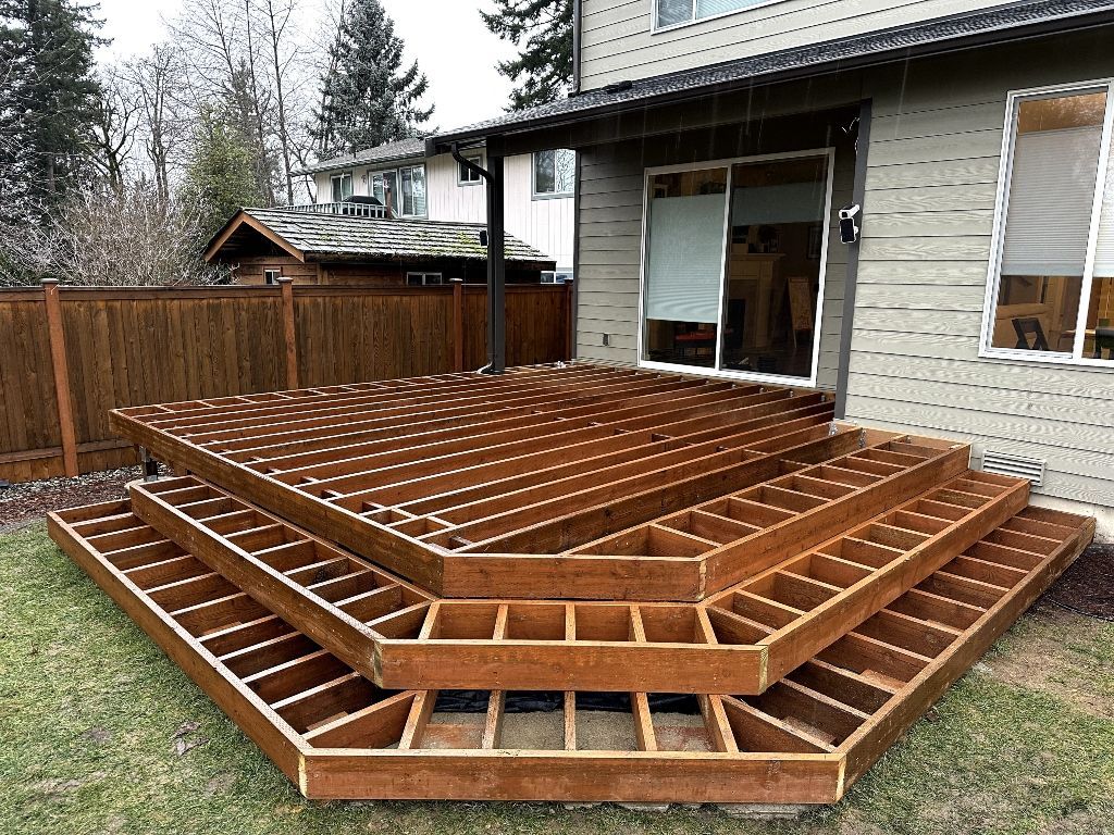 Wooden deck frame under construction beside a house, with exposed joists and steps.