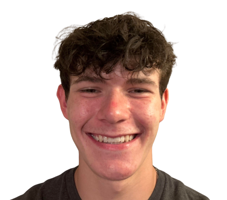 Young man with curly brown hair smiles broadly, appearing happy. He's wearing a gray shirt, against a white background.