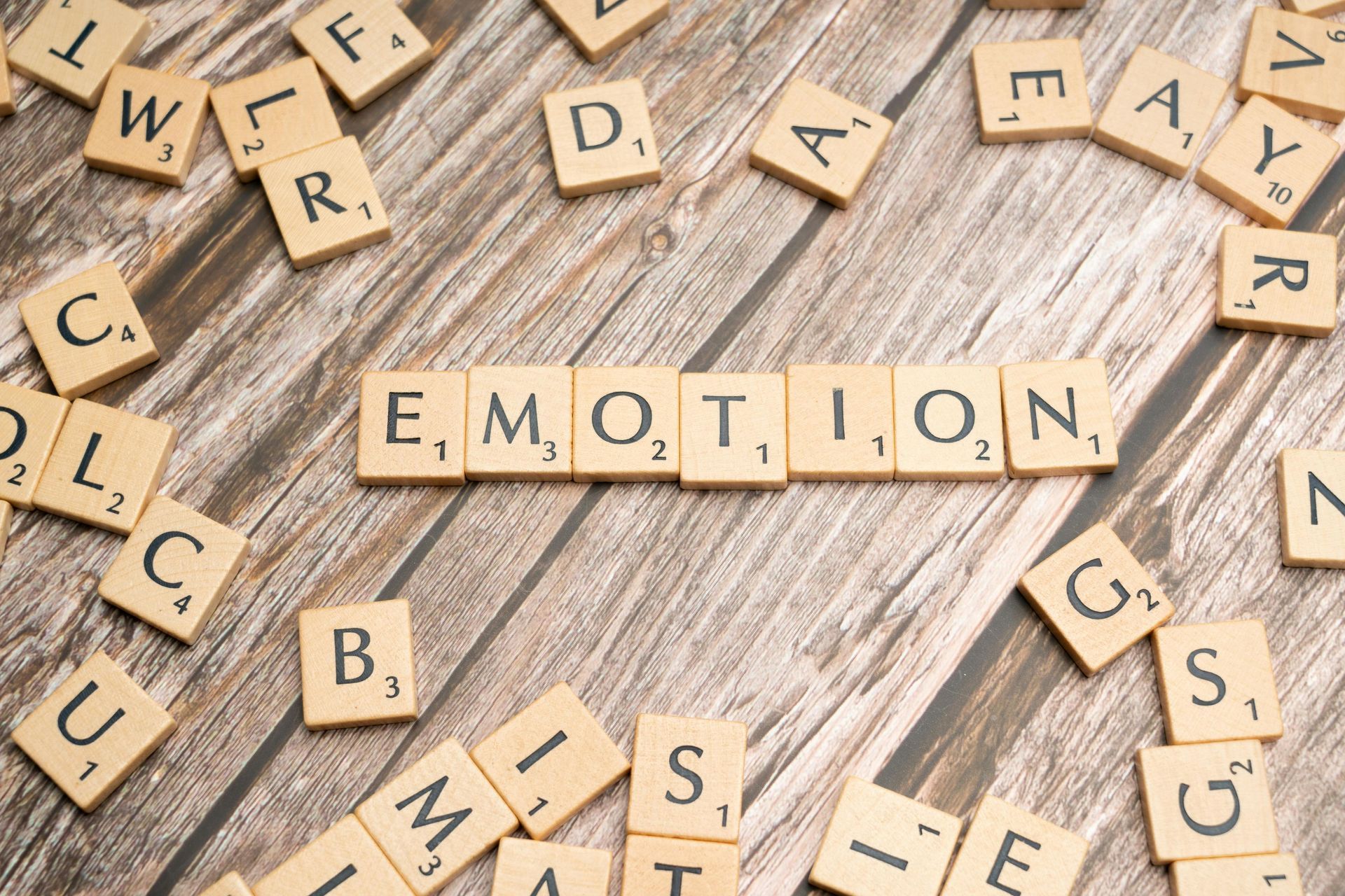 Scrabble tiles spelling “EMOTION” on a wooden surface, with scattered letter tiles around them