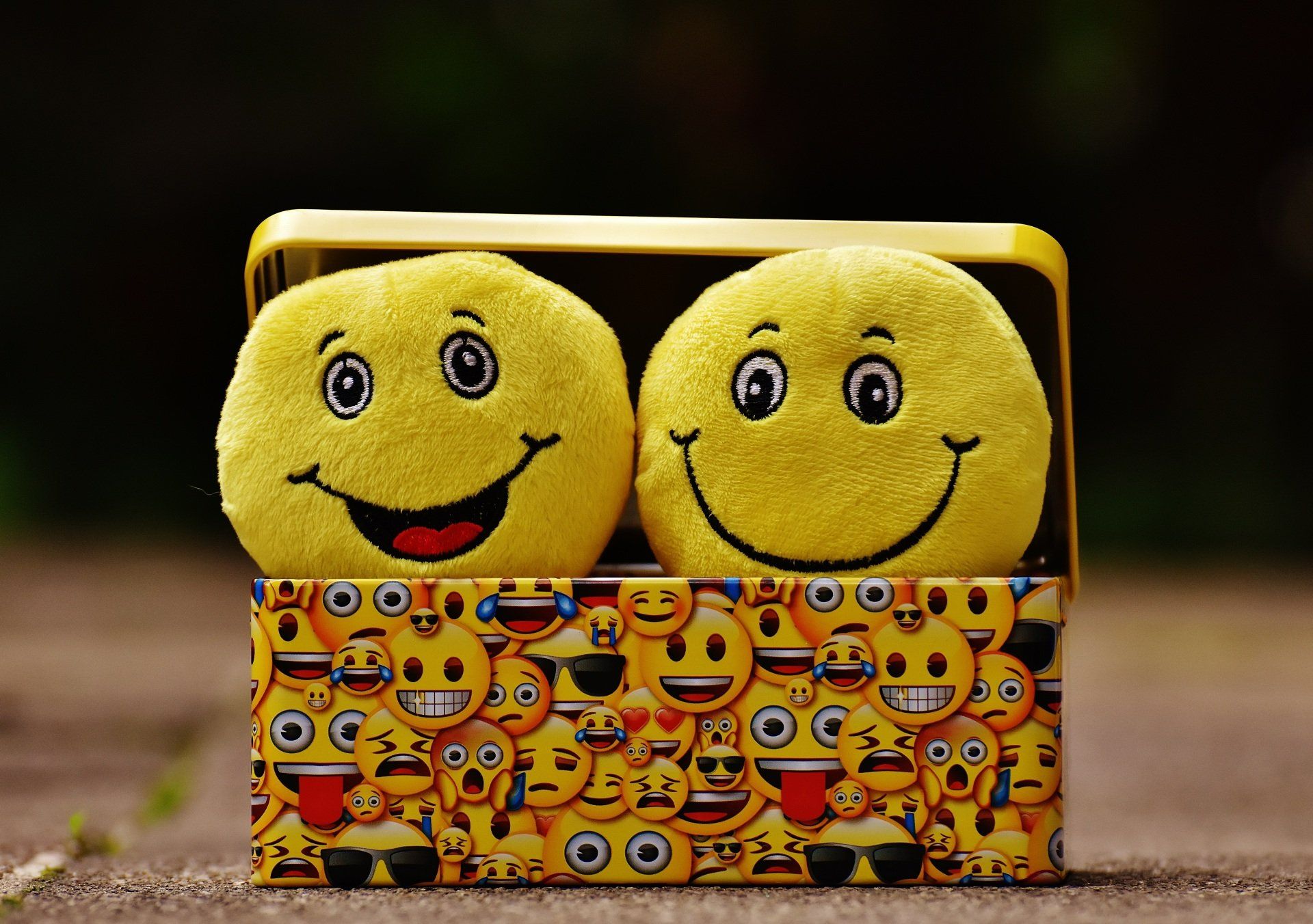 Two yellow smiley plush balls in a small box with a colorful emoji pattern.