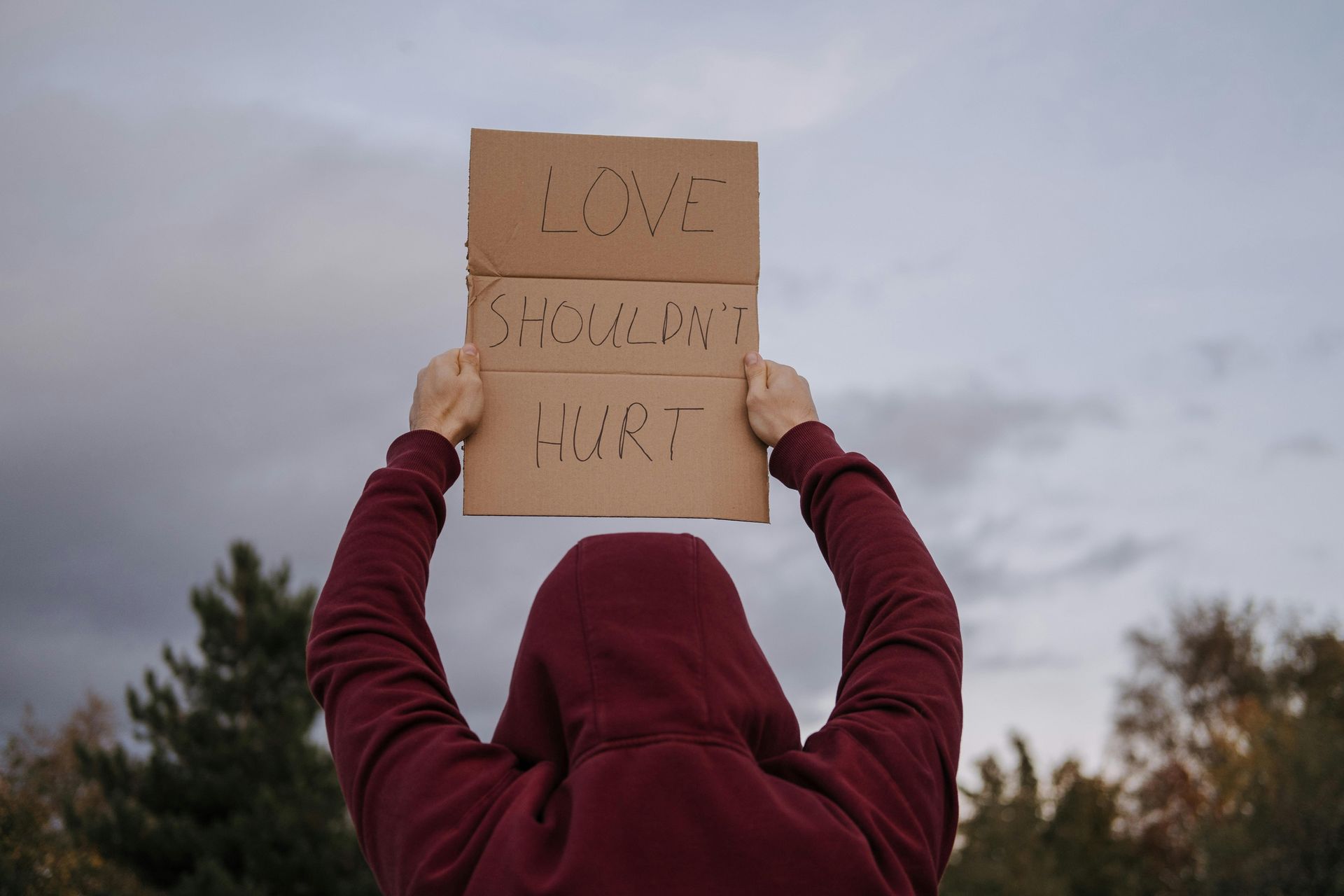 Person in a maroon hoodie holding a cardboard sign reading “LOVE YOUR NEIGHBOR” overhead outdoors