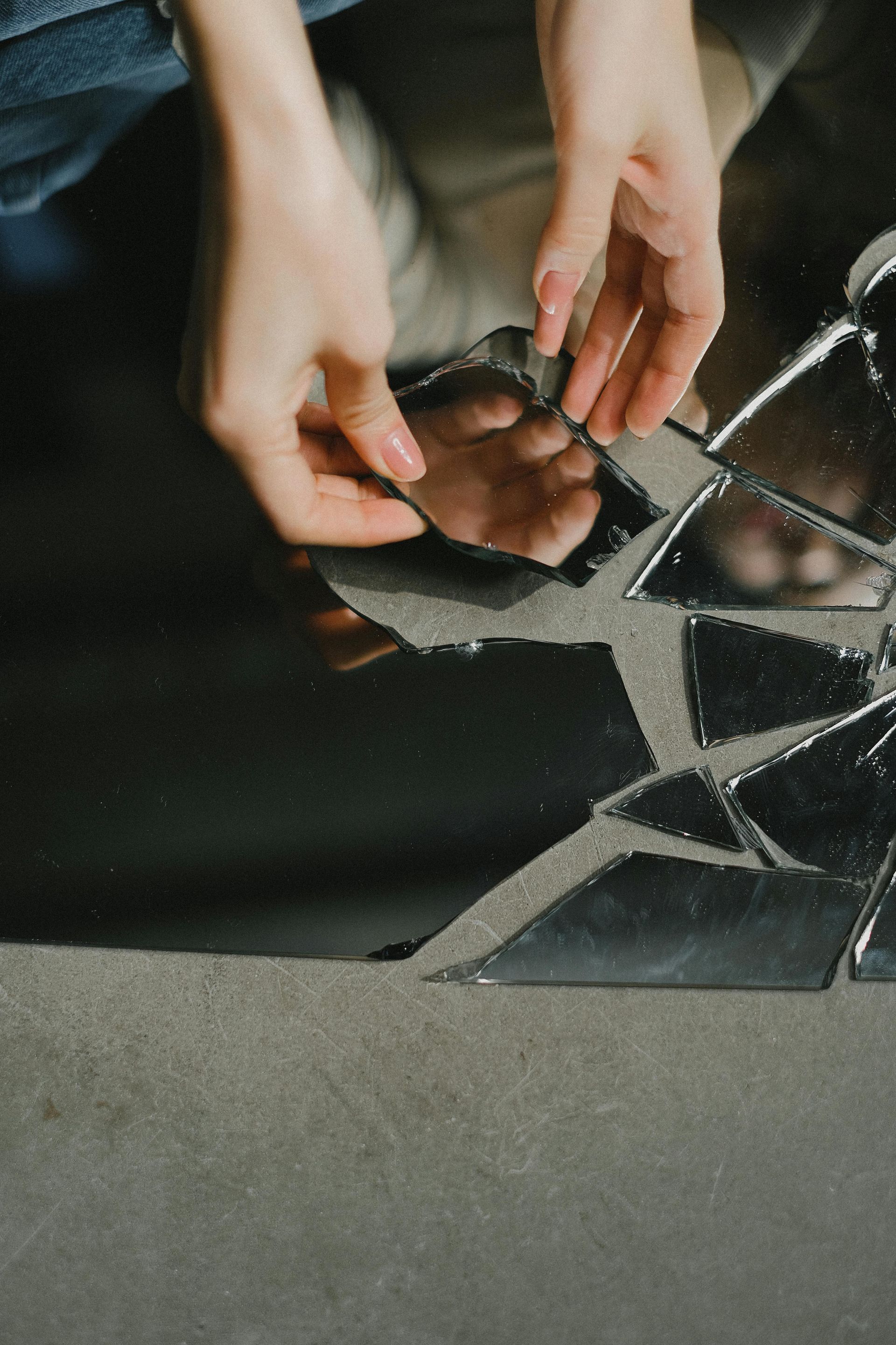Hands lifting a shattered mirror with a black background and reflections