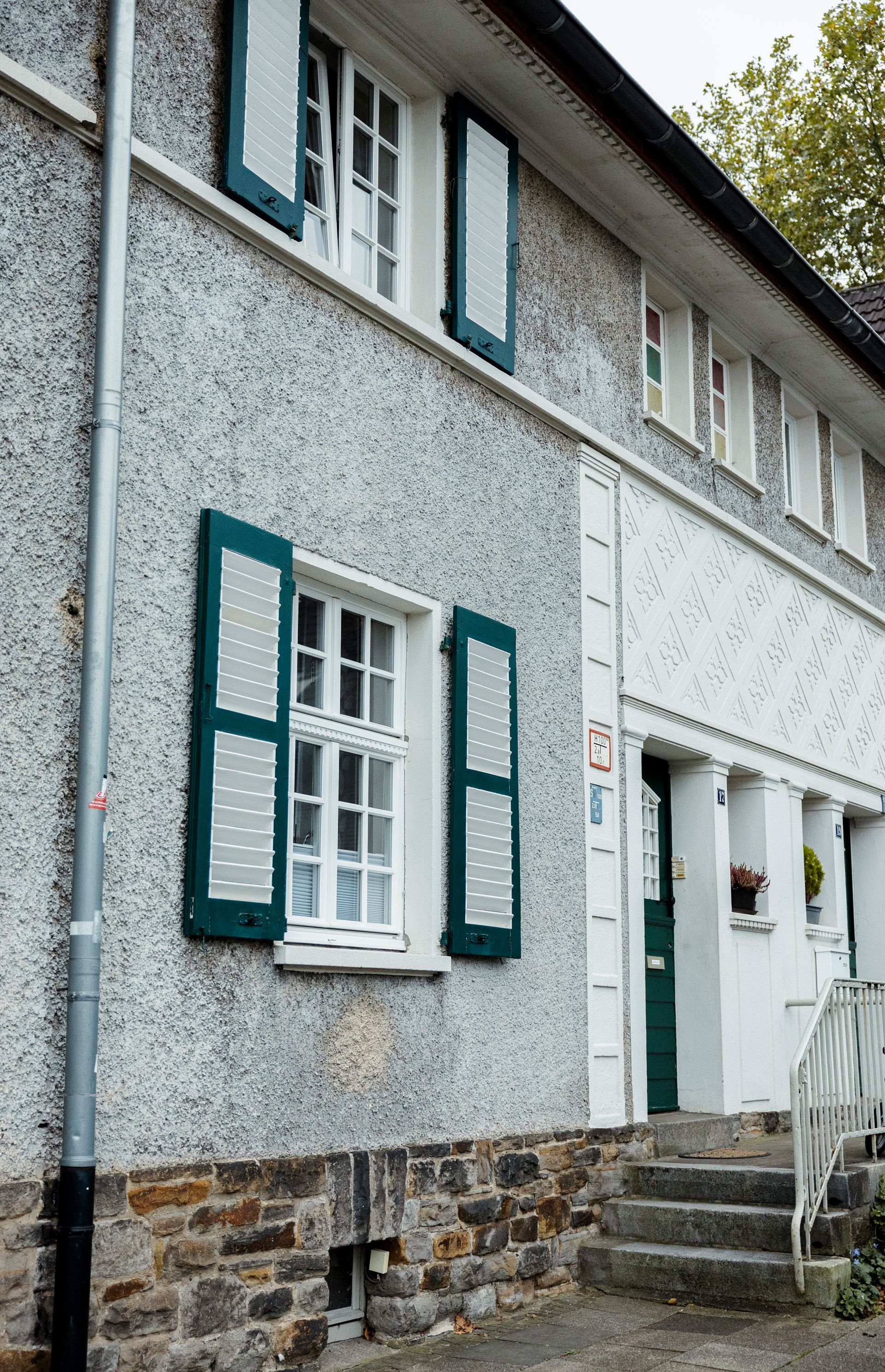 Graues Stuckhaus mit grünen Fensterläden und weißen Zierelementen, Stufen führen zur Haustür.