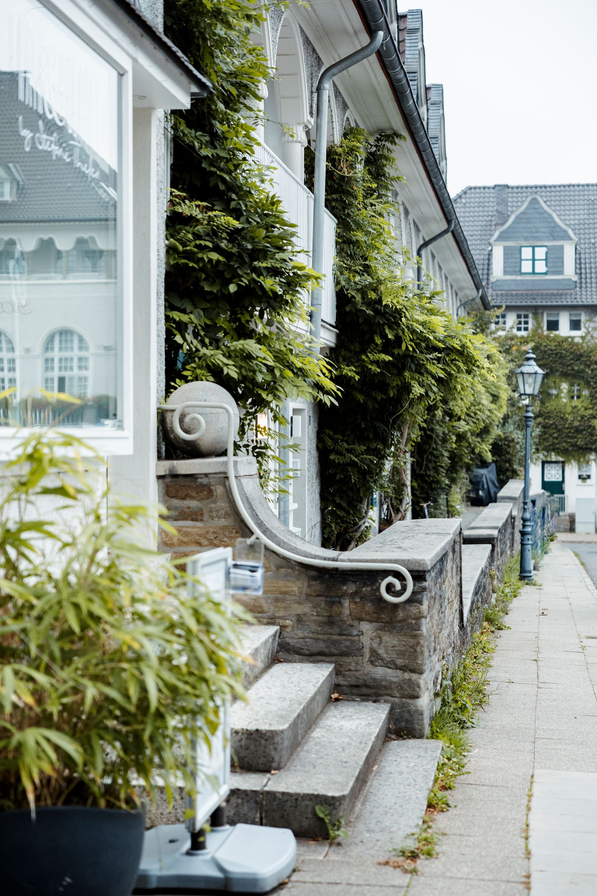 Steintreppen führen zu einem Gebäude mit einem weißen, mit grünen Ranken bewachsenen Balkon, neben einem Bürgersteig.