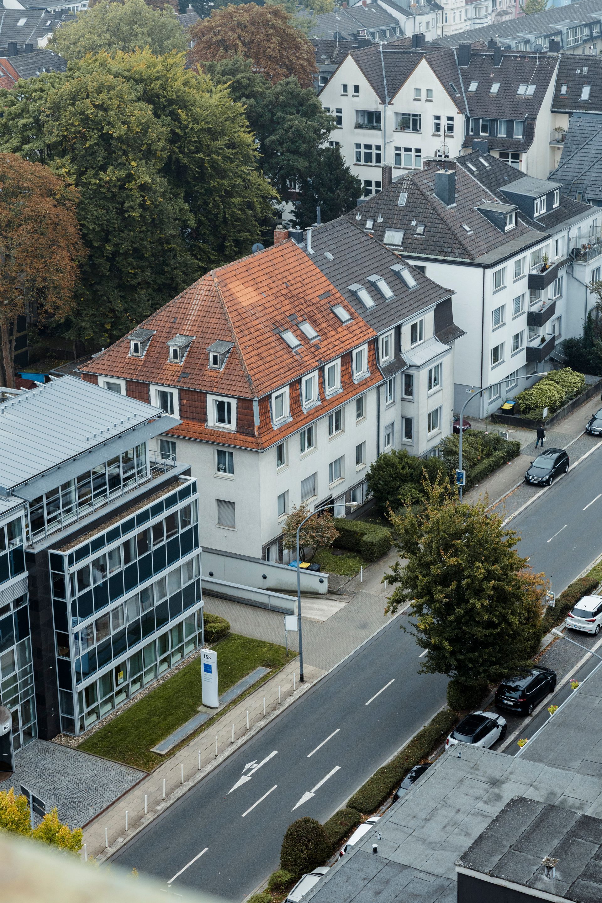 Blick auf Gebäude und Straße, teilweise mit Bäumen und geparkten Autos, bedeckter Himmel.