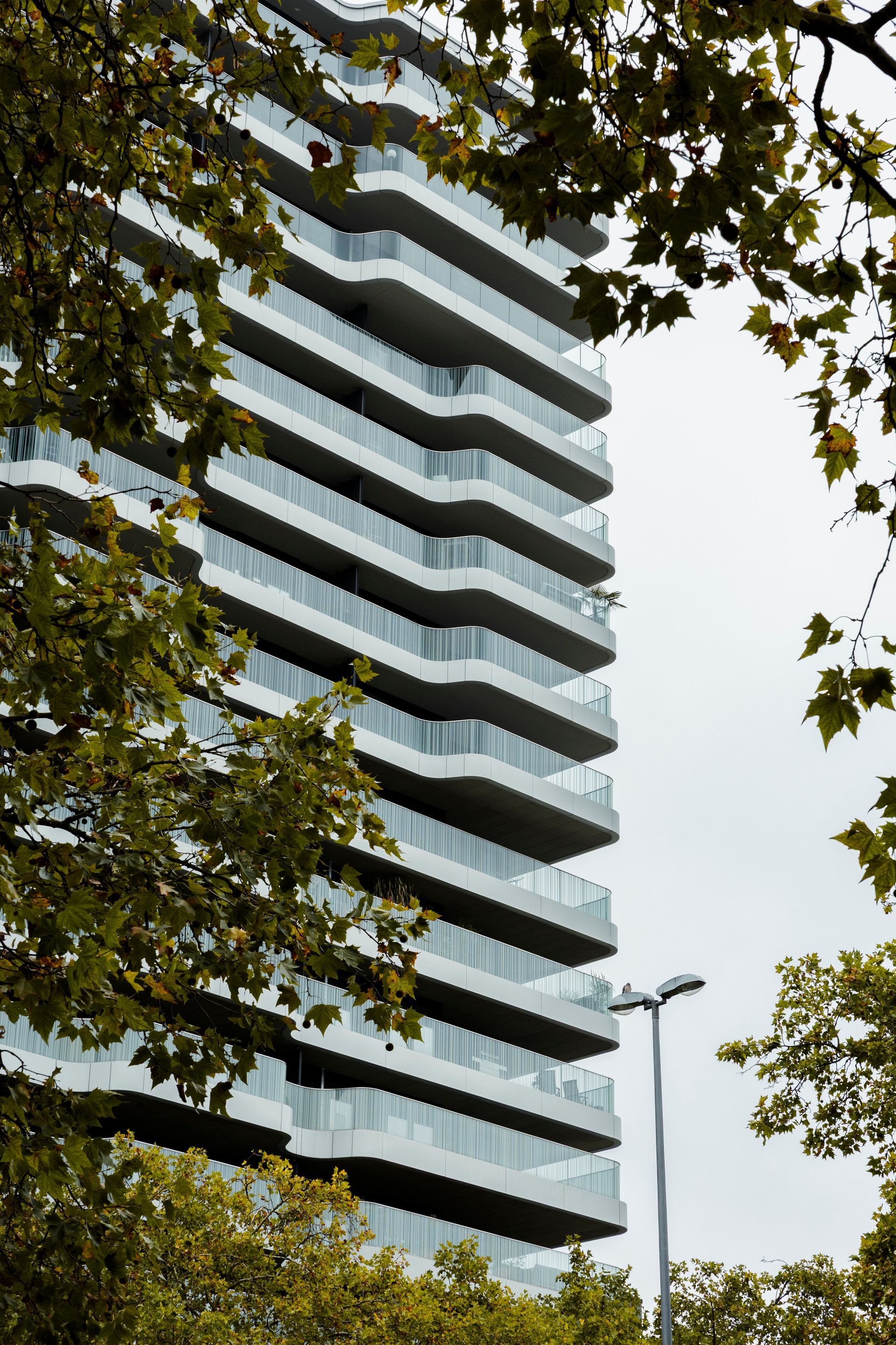 Modernes weißes Hochhaus mit zickzackförmigen Balkonen, eingerahmt von grünen Baumzweigen an einem bewölkten Tag.