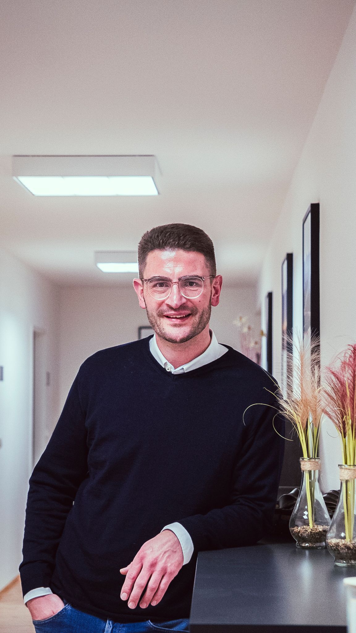 Ein Mann mit Brille und dunkelblauem Pullover lehnt an einer dunklen Theke in einem Flur und lächelt leicht.