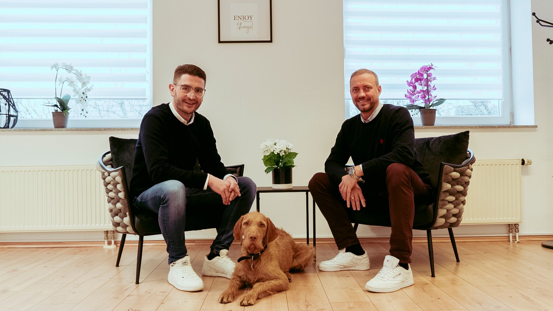 Zwei Männer und ein Hund sitzen lächelnd in einem Haus. Der eine trägt Jeans, der andere eine braune Hose.