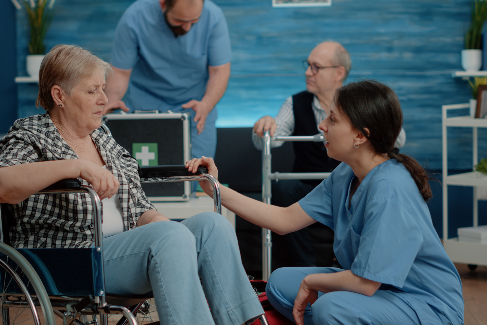 Woman in wheelchair with nurse, man with walker, another with open first aid kit, blue walls.