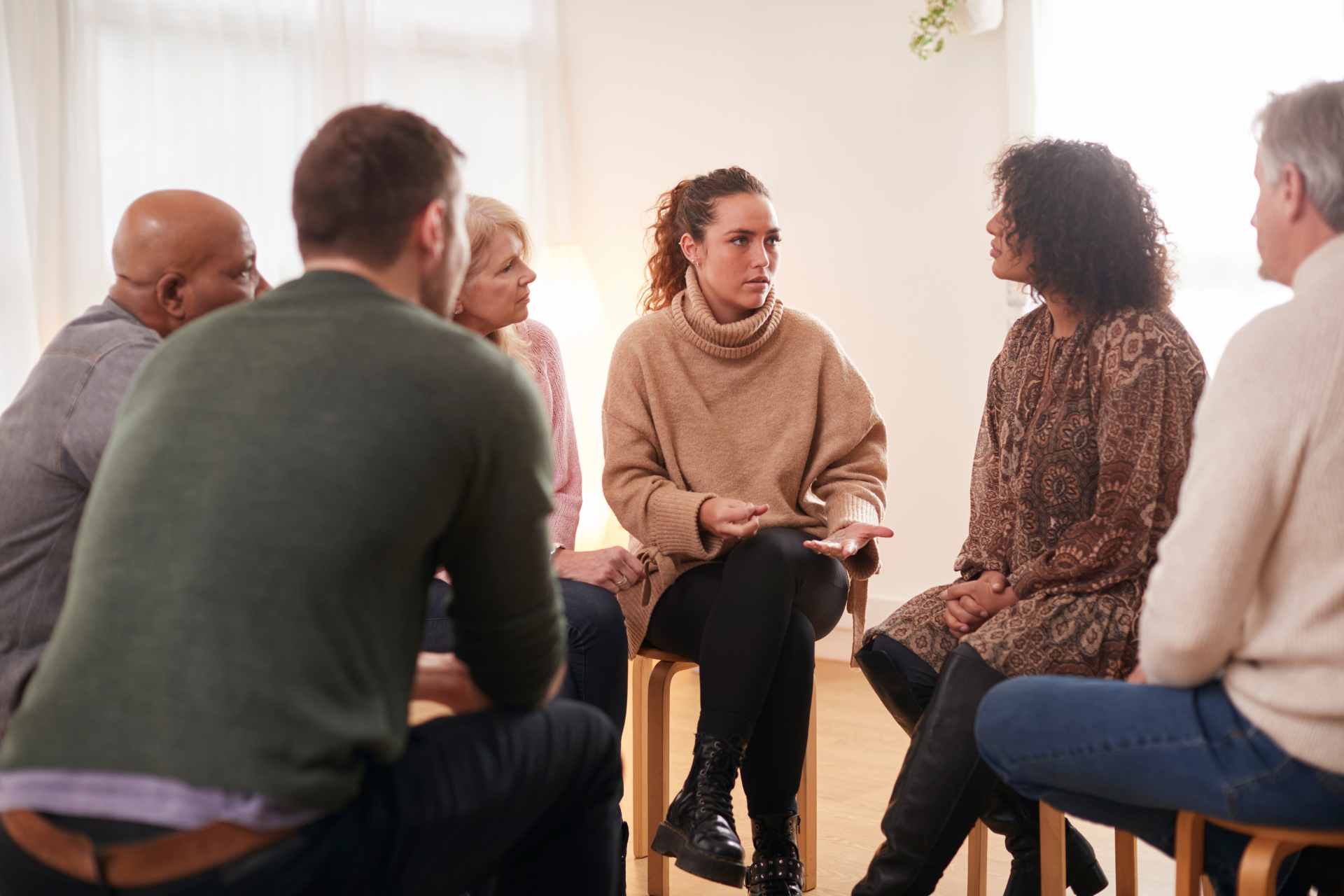 Group of people in a circle, talking in a room. Woman in center speaks while others listen.