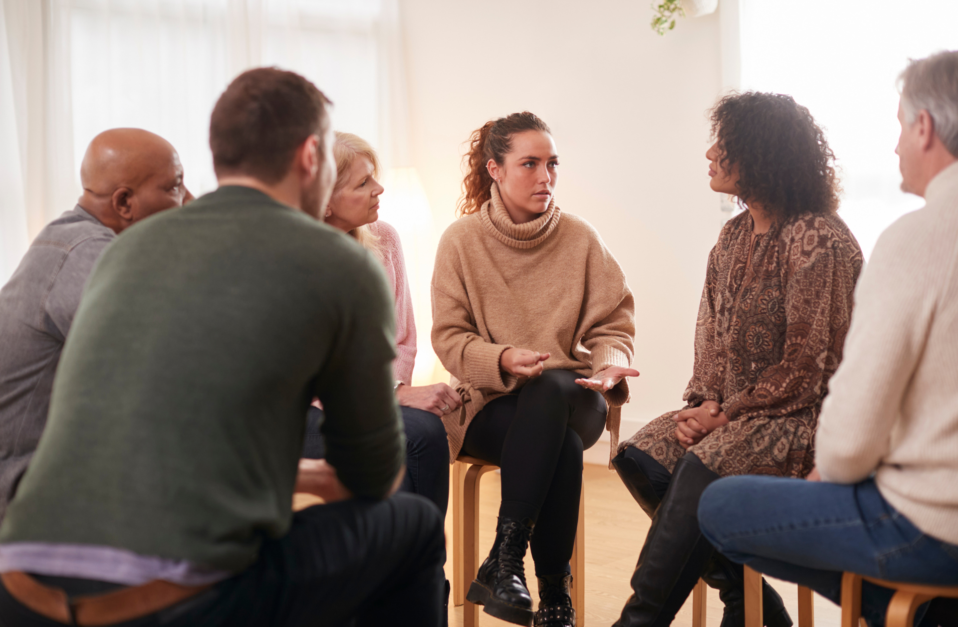 Group therapy session: People sit in a circle, conversing. A woman in a sweater is speaking.