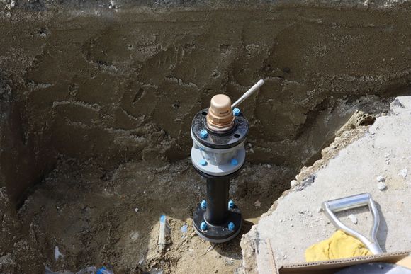 Black pipe with blue bolts, brass top, in a dirt trench, near concrete, with a shovel. Black pipe with blue bolts, brass top, in a dirt trench, near concrete, with a shovel.