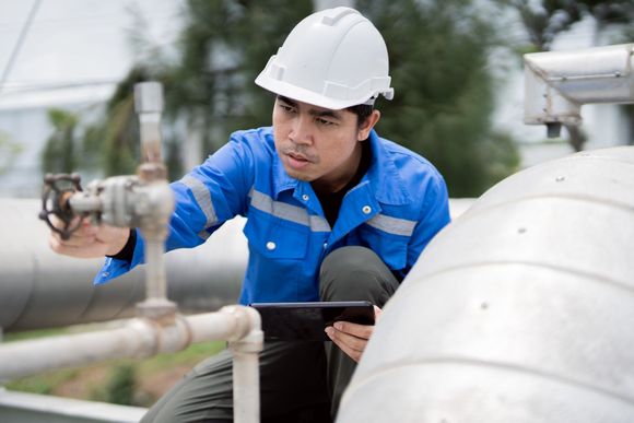 Worker in blue coveralls and hard hat inspecting pipes, holding a tablet. Worker in blue coveralls and hard hat inspecting pipes, holding a tablet.