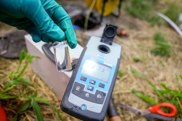 Gloved hand holding a water sample vial near a digital water quality meter outdoors; grass background. Gloved hand holding a water sample vial near a digital water quality meter outdoors; grass background.