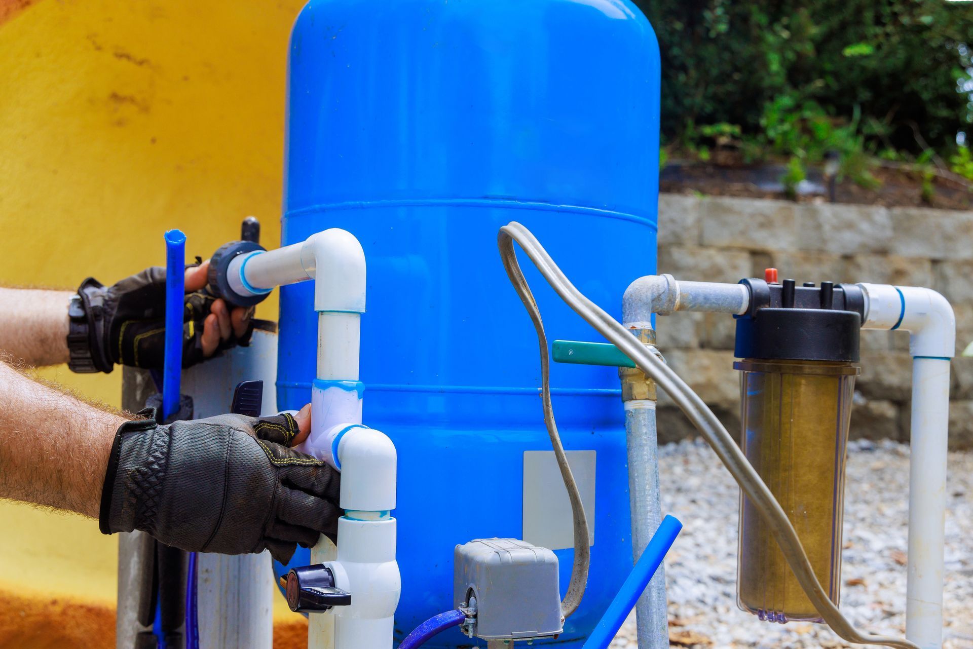 Person in gloves connecting white PVC pipes to a blue water tank and filter. Outdoor setting.
