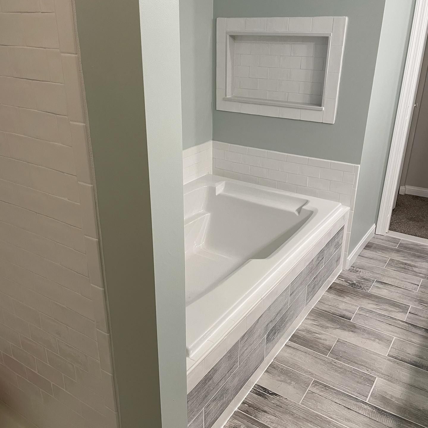 Bathroom with white tub, light blue walls, gray tile surround, and a built-in shelf.