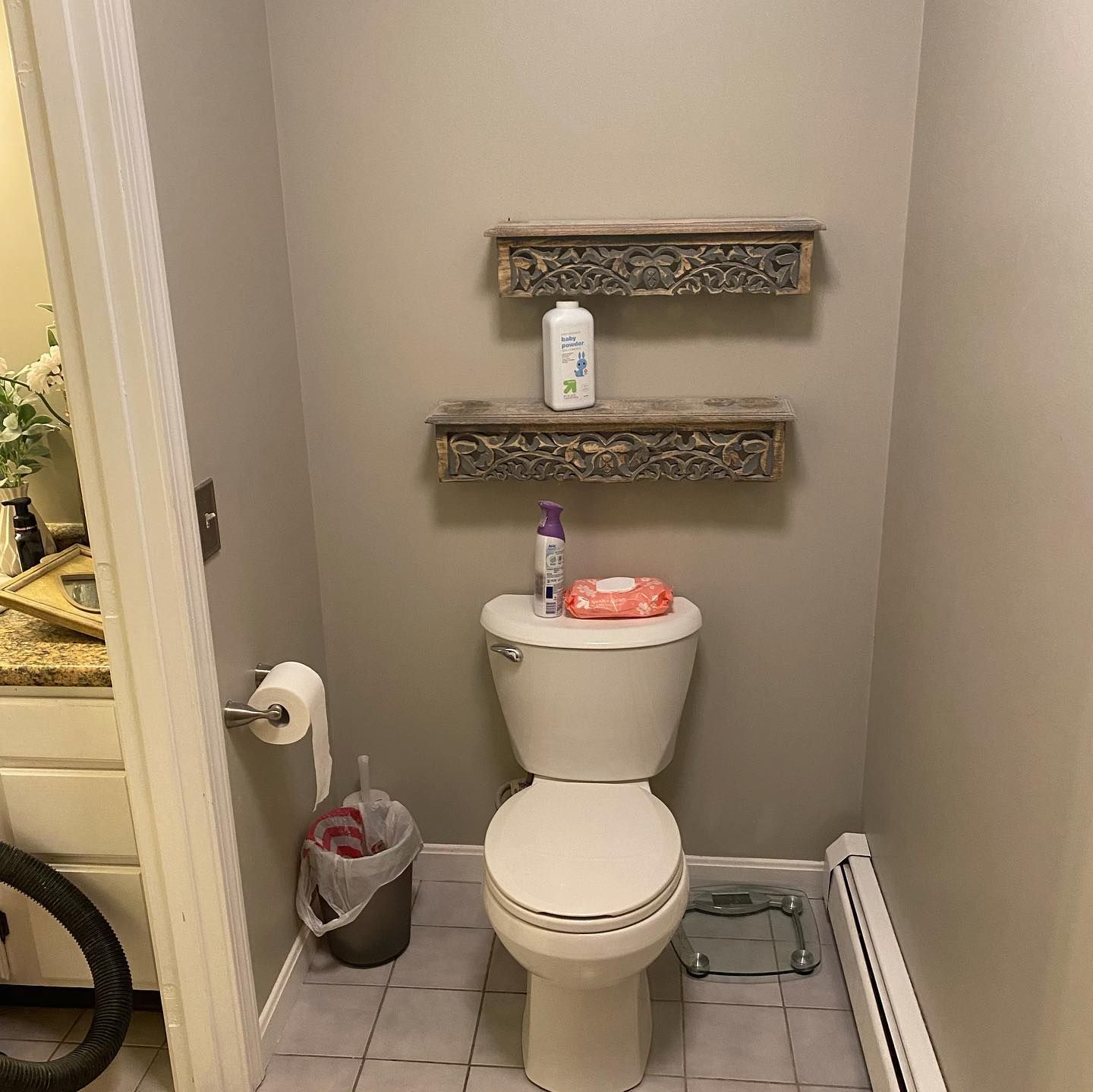 Small bathroom with toilet, shelves, and gray walls. Includes a toilet paper roll, trash can, and a scale.