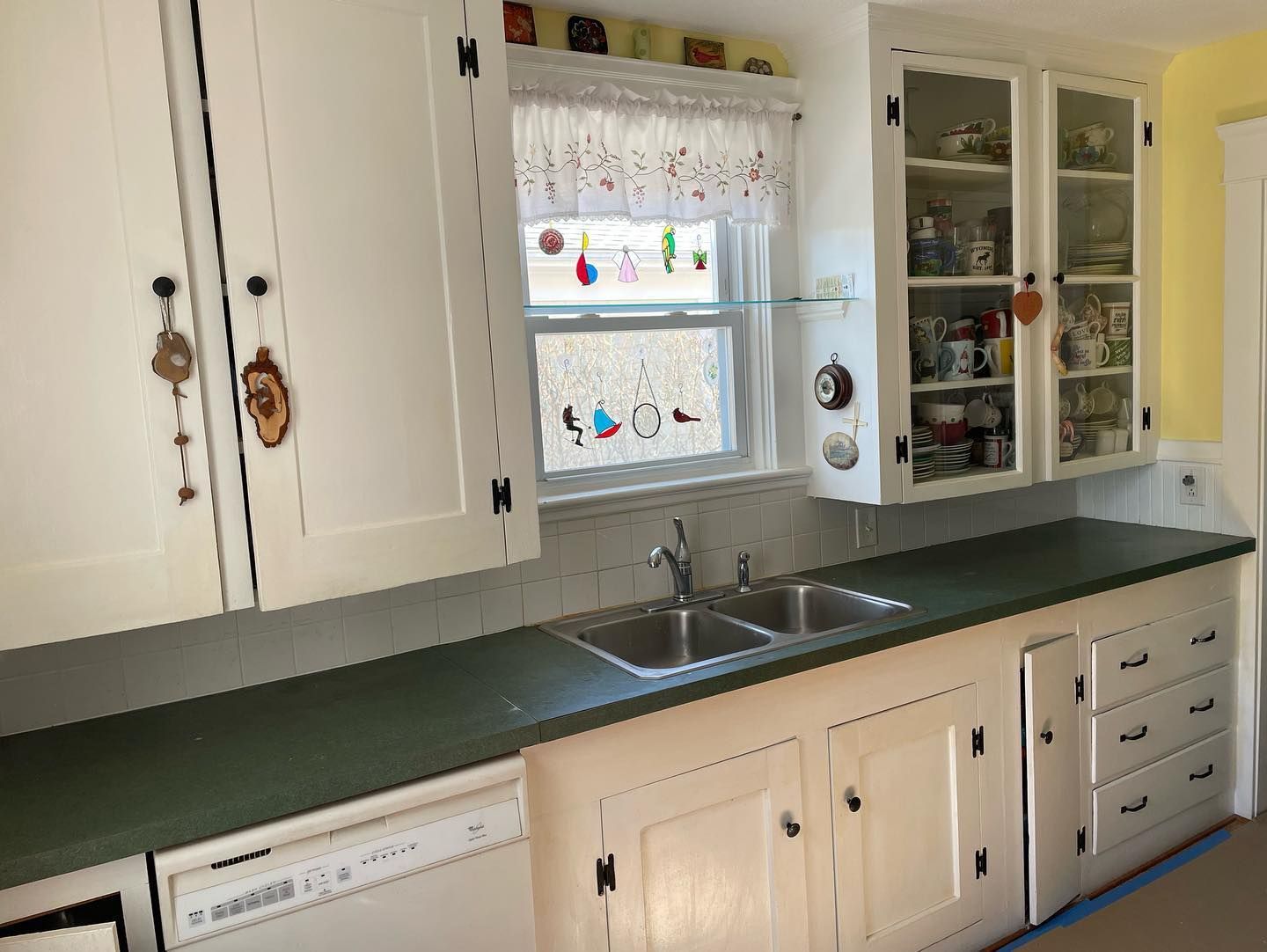 White kitchen cabinets with dark green countertop, double sink, and a window with colorful decorations.
