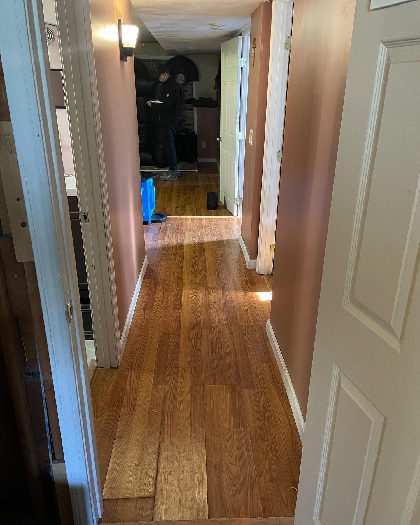 Narrow hallway with wood floor, pink walls, and open doors. A person stands at the end of the hallway.