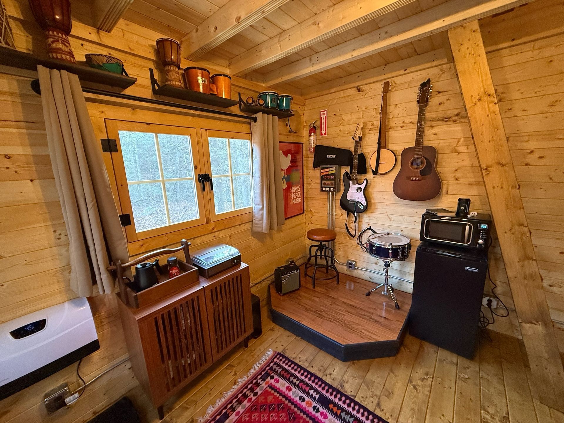 Cozy log cabin interior with wood walls, window, small desk, guitar, and chair on a colorful rug.