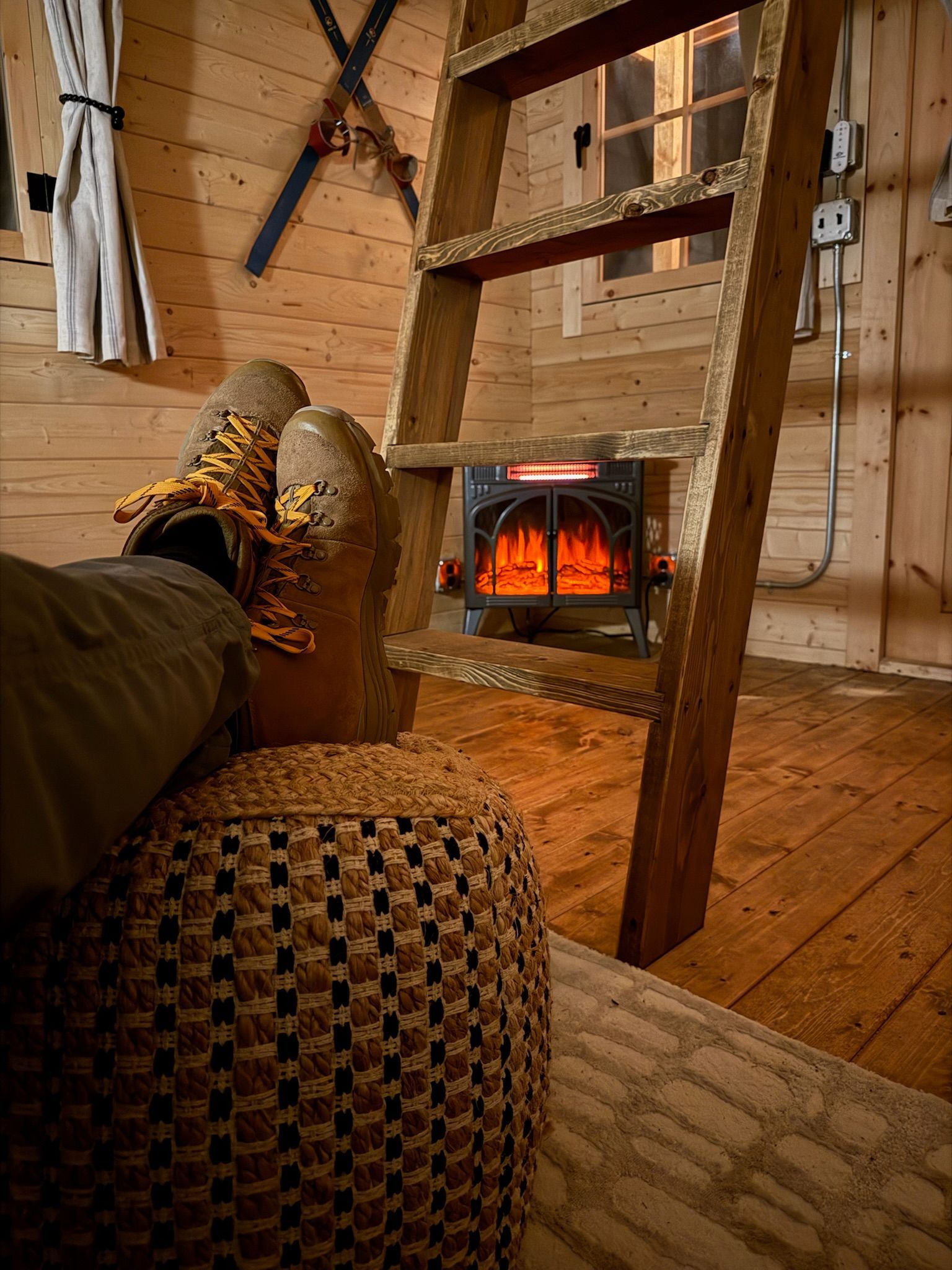 Cozy cabin interior with a lit wood stove, boots on a woven pouf, and a rustic wooden ladder.
