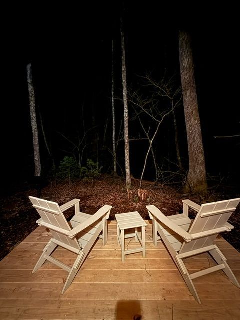 Two wooden chairs and a small stool on a deck at night, facing a dark wooded area.