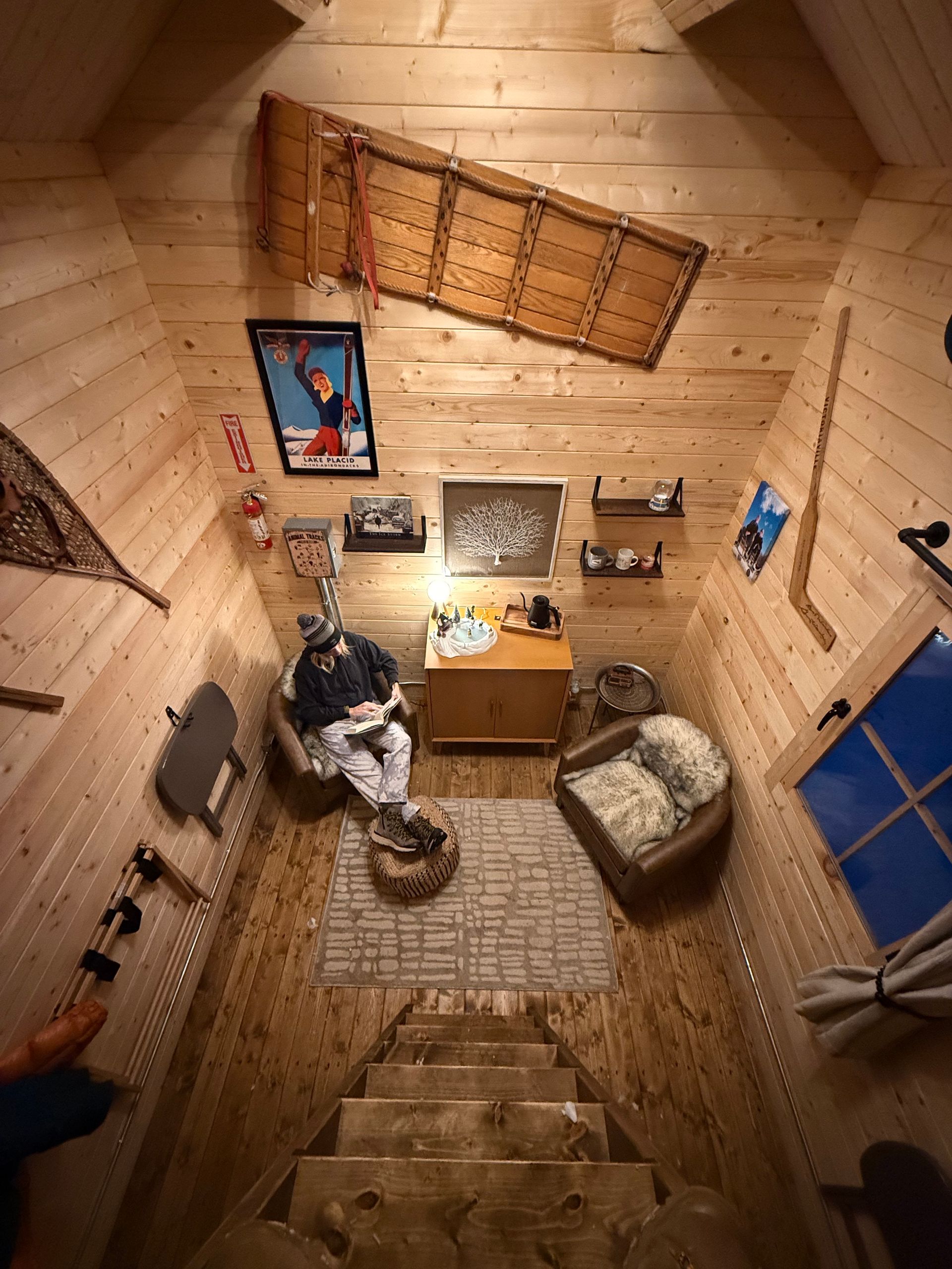 Top-down view of a cozy wooden staircase room with a desk, chair, rug, wall art, and a person sitting below.