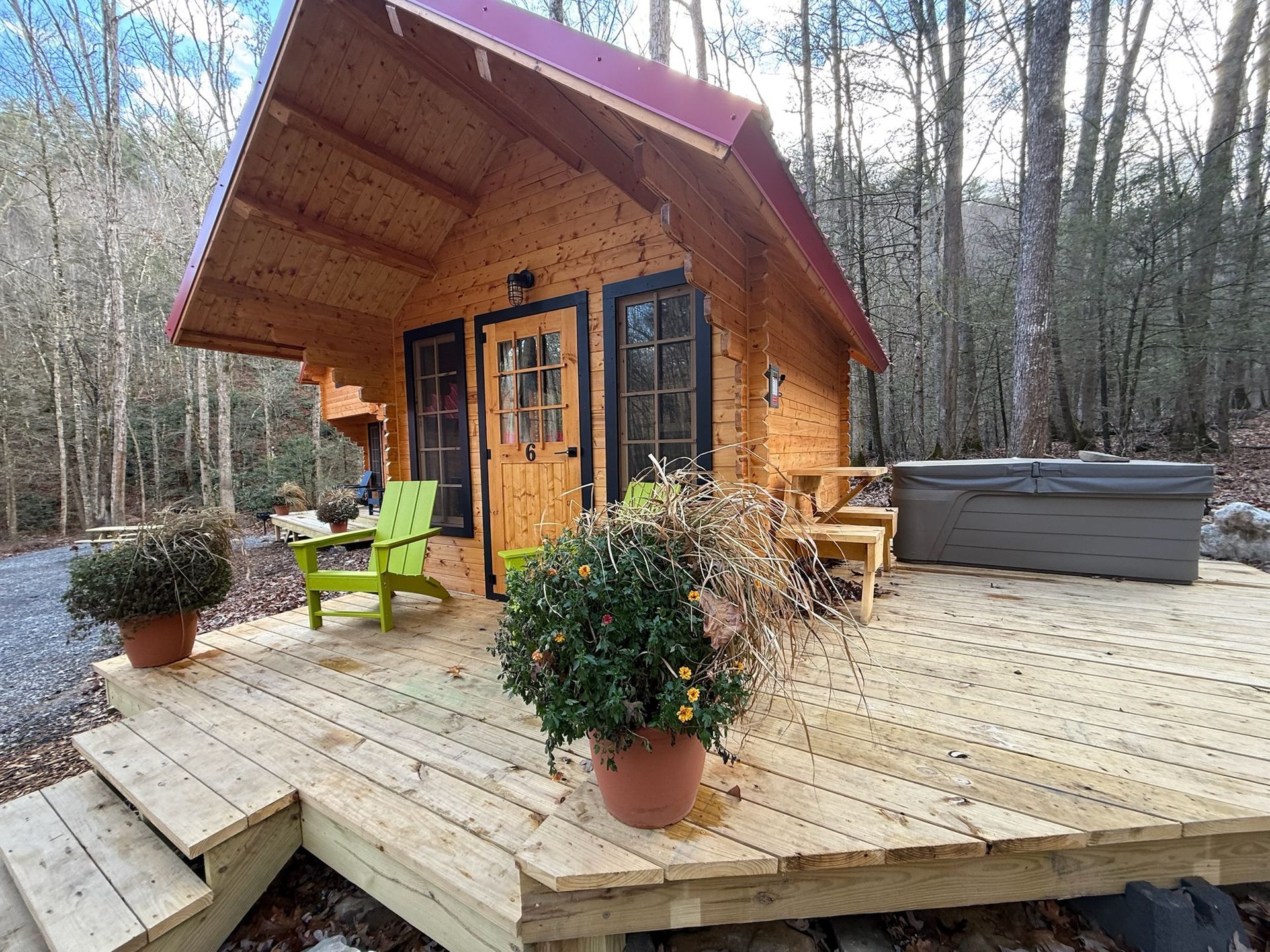Small wooden cabin with deck, green chairs, potted plants, and a hot tub in a wooded setting