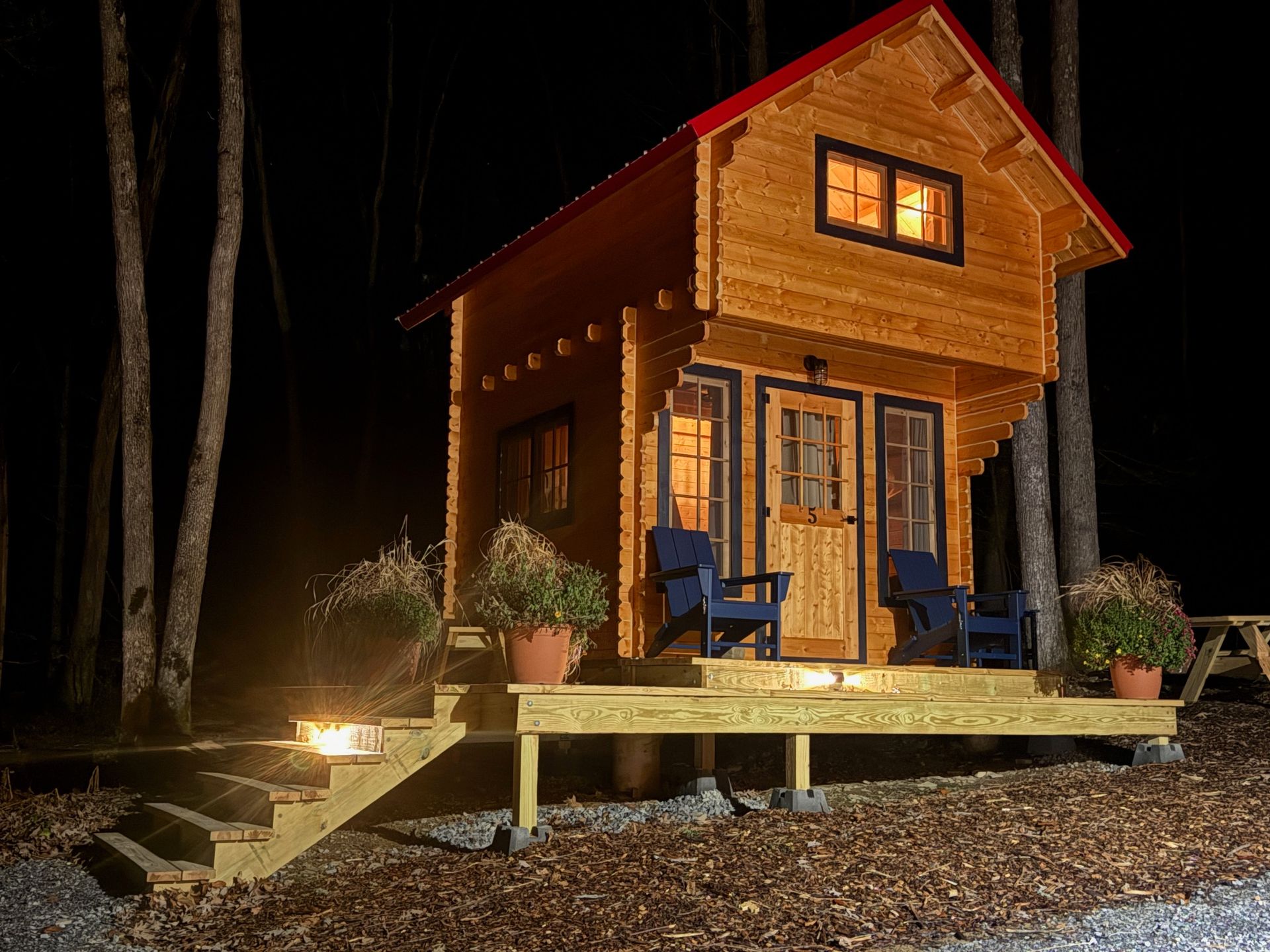 Small lit wooden cabin at night with a porch, steps, chairs, and potted plants in a wooded setting