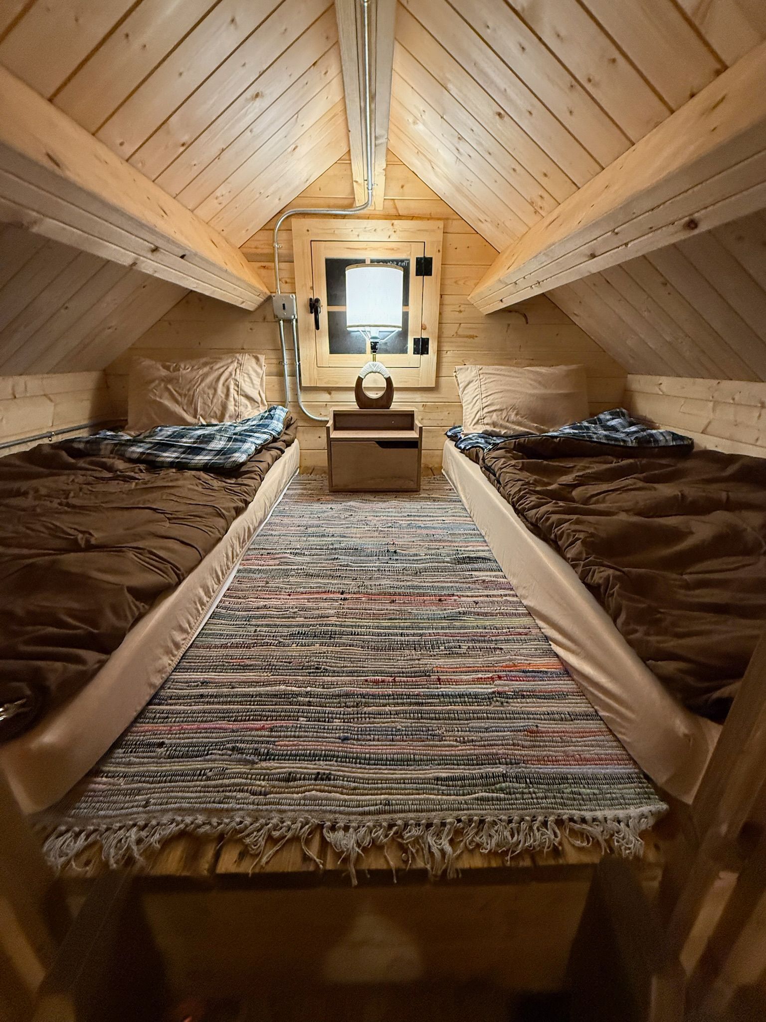 Cozy attic bedroom with twin beds, patterned rug, and a small window at the end.