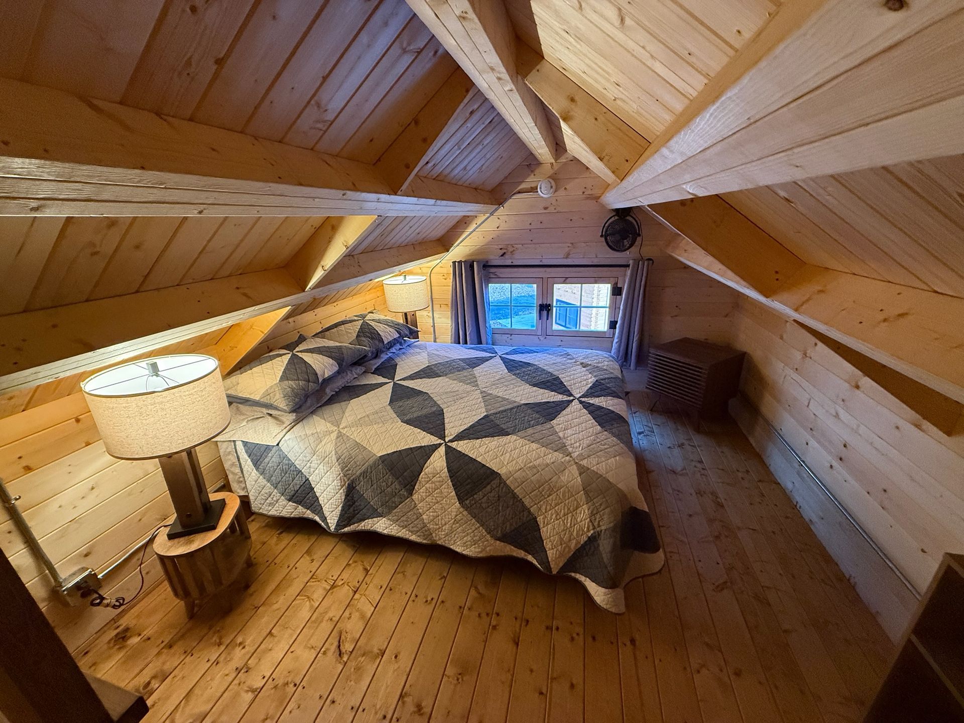 Cozy attic bedroom with sloped wood-paneled ceiling, quilted bed, lamp, and small window seat.