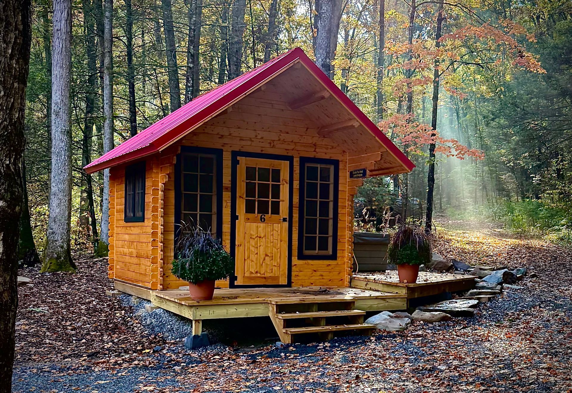 Small wooden cabin with a red roof in a forest clearing, surrounded by autumn leaves.