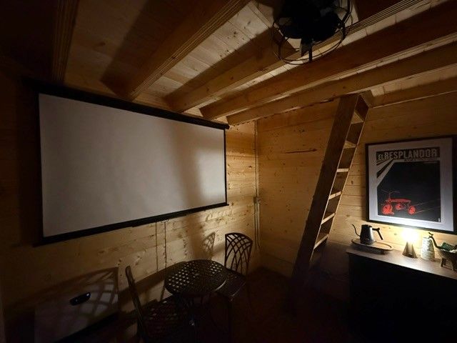 Cozy wood-paneled attic with projector screen, ladder, small table, and lit desk beside a framed poster