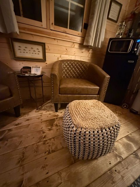 Cozy rustic room with a leather chair, woven ottoman, side table, and wood-paneled walls.