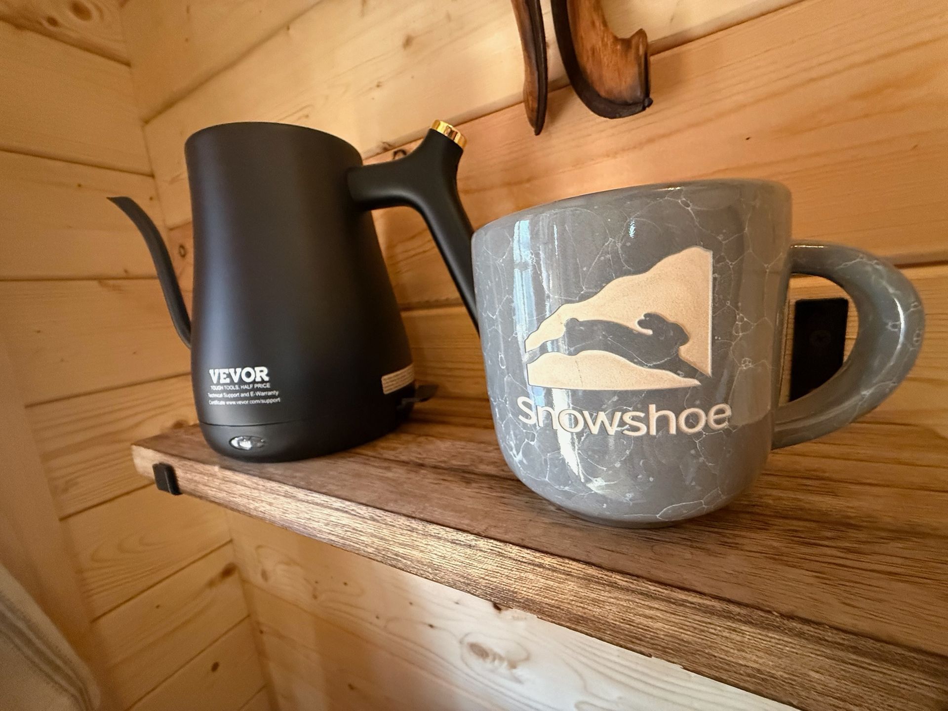Black electric kettle and gray Snowshoe mug on a wooden shelf in a rustic cabin interior