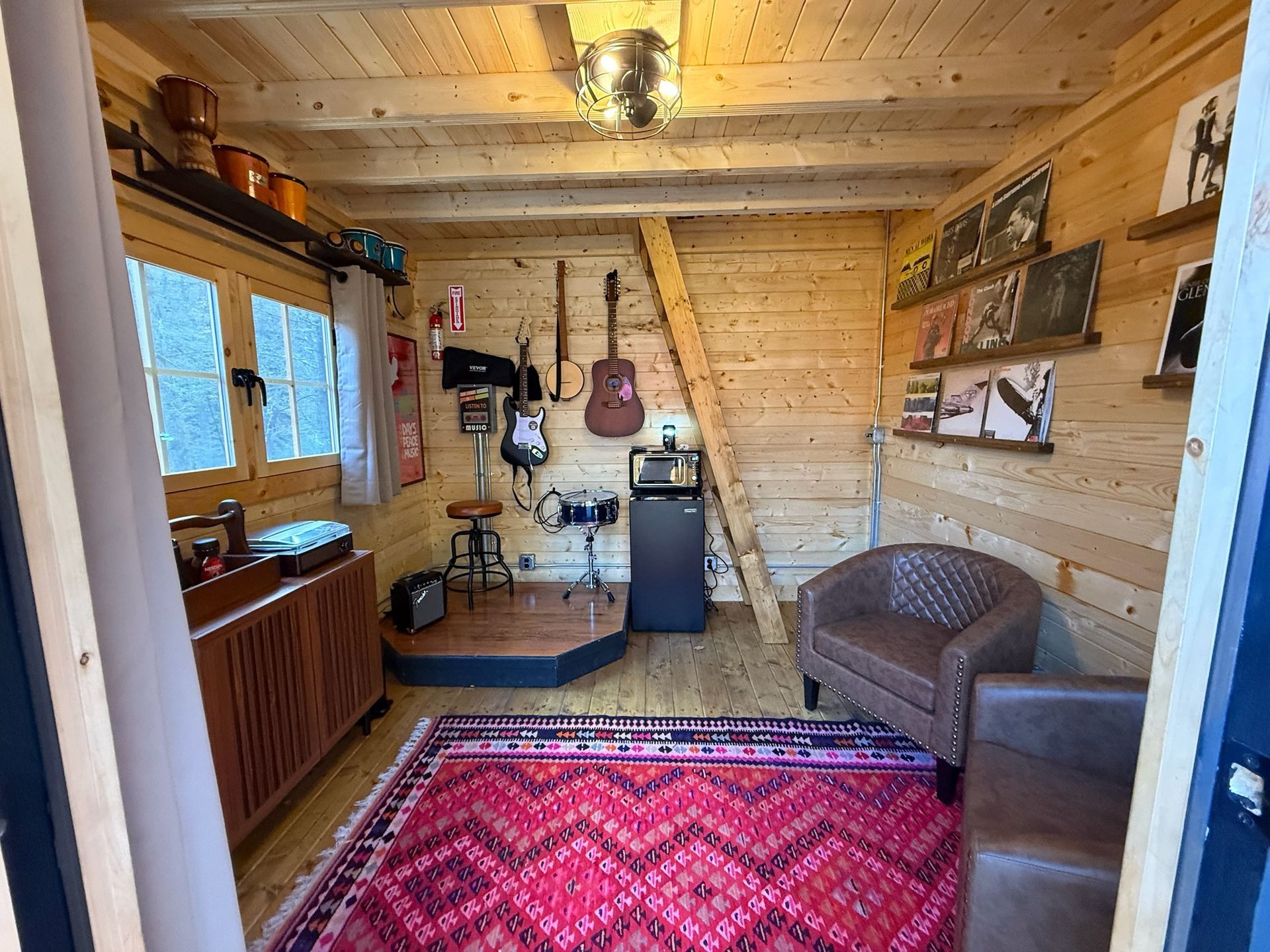 Cozy wood-paneled room with red rug, chair, desk, window, and ladder leaning against the wall