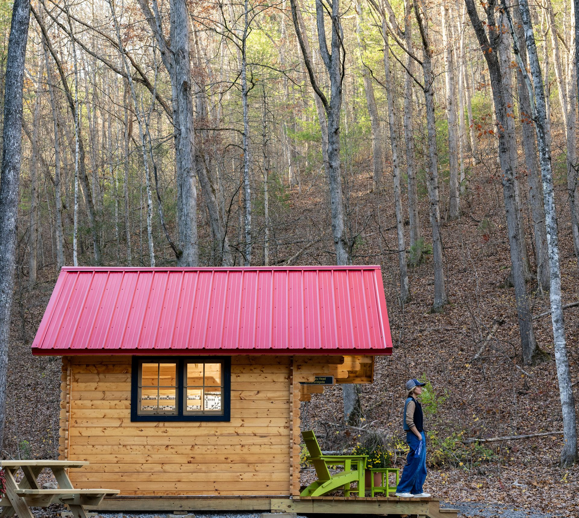 Small wooden cabin with a red roof in a forest, with a person standing outside near the door.
