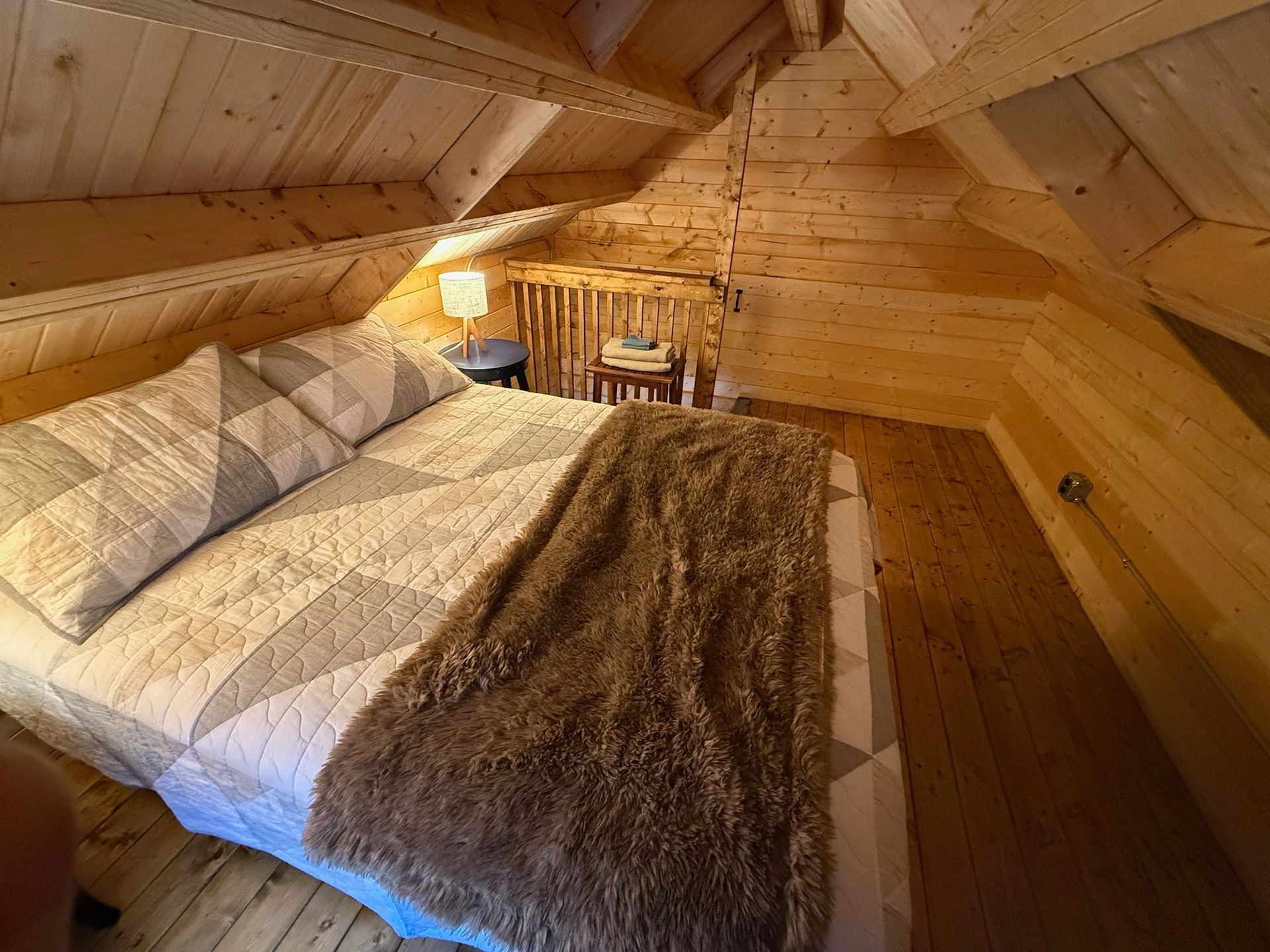 Cozy attic bedroom with a wooden vaulted ceiling, bed, fur throw, and small table lit by warm light