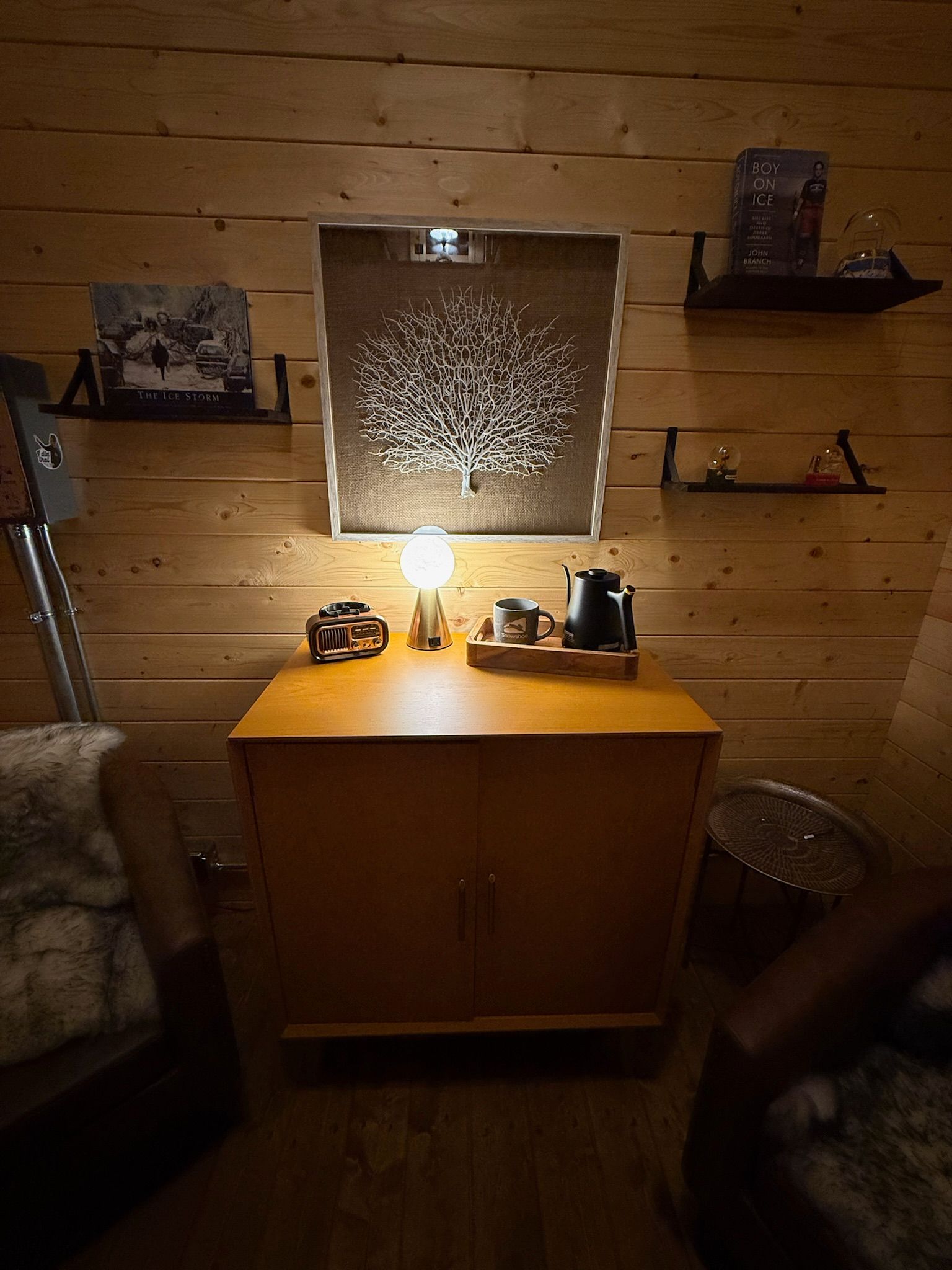 Warm wooden room with a glowing table lamp, cabinet, wall art, and shelves with small decor pieces.