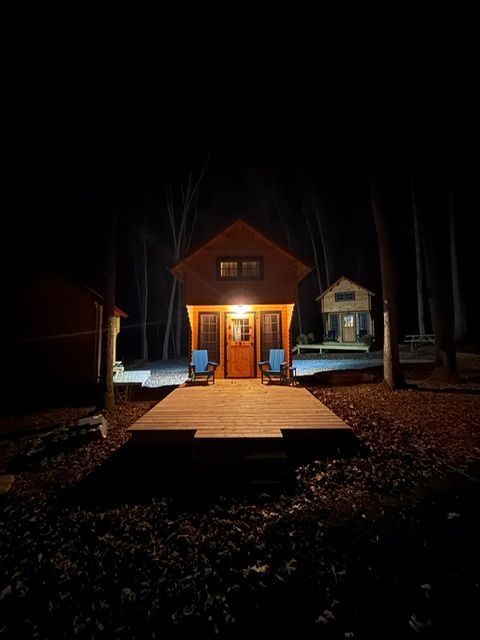 Small lit cabin at night with a wooden deck and steps, surrounded by dark trees and fallen leaves.
