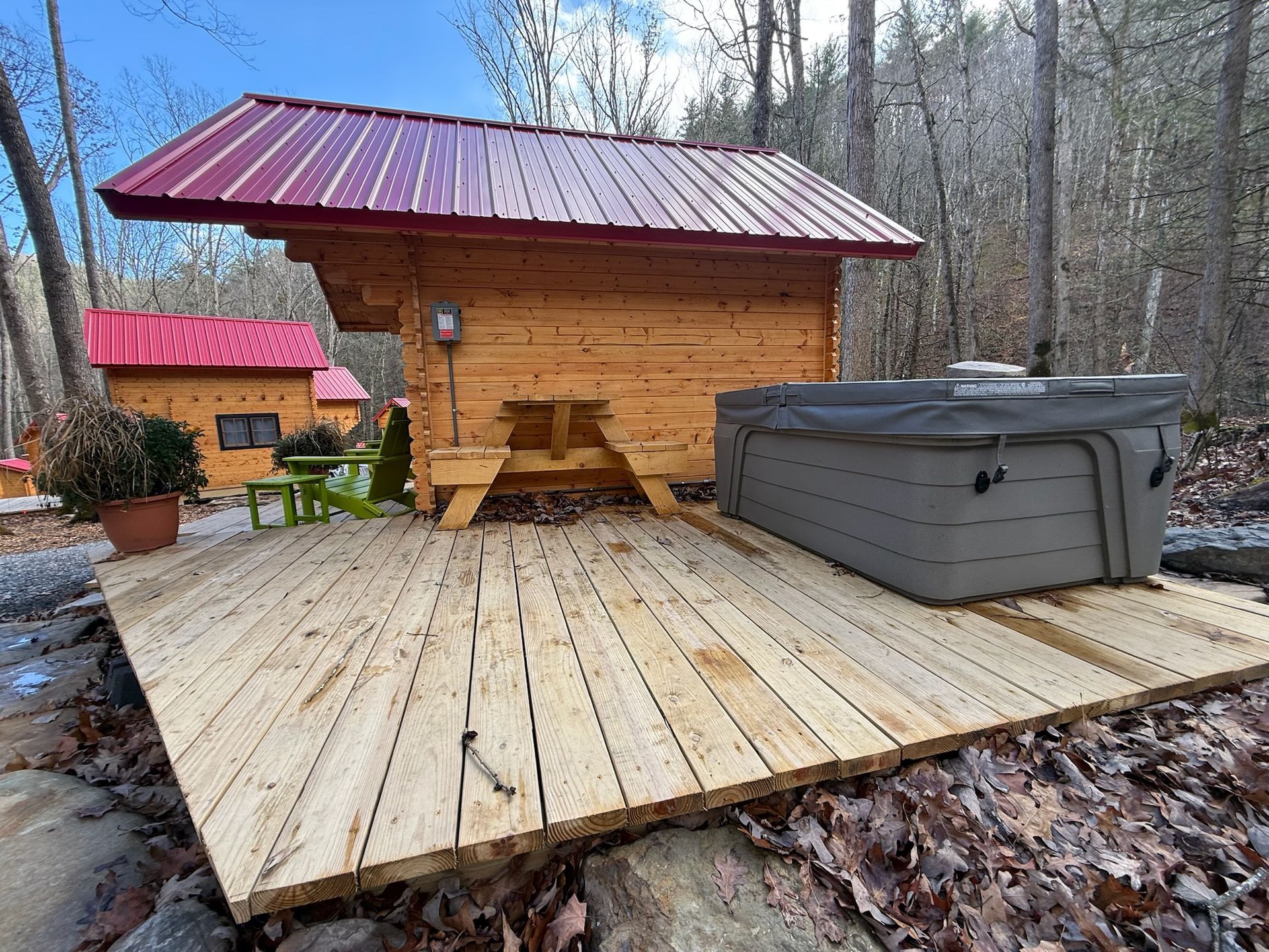 Wooden cabin deck with hot tub and picnic table in a wooded backyard, red metal roof overhead