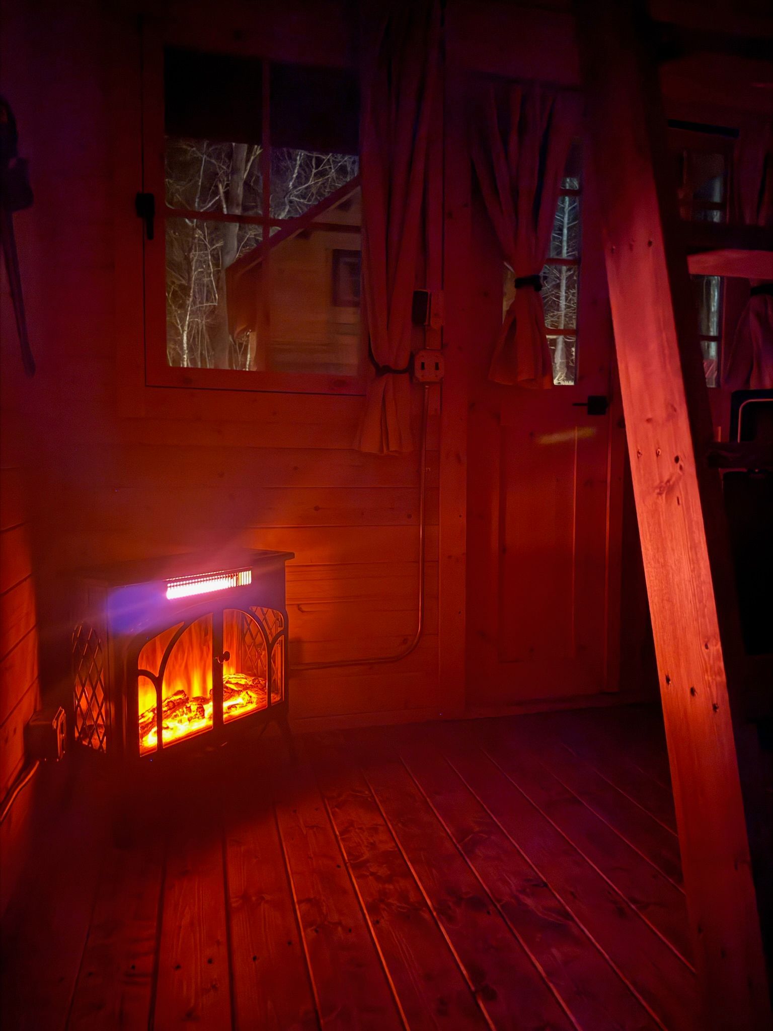 Dim wooden room lit by a glowing fireplace, with red curtains and a window above.
