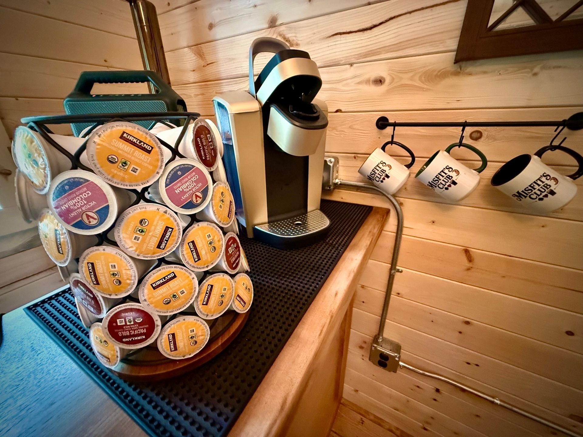Single-serve coffee pod organizer beside a coffee maker on a wooden counter, with mugs hanging on the wall.