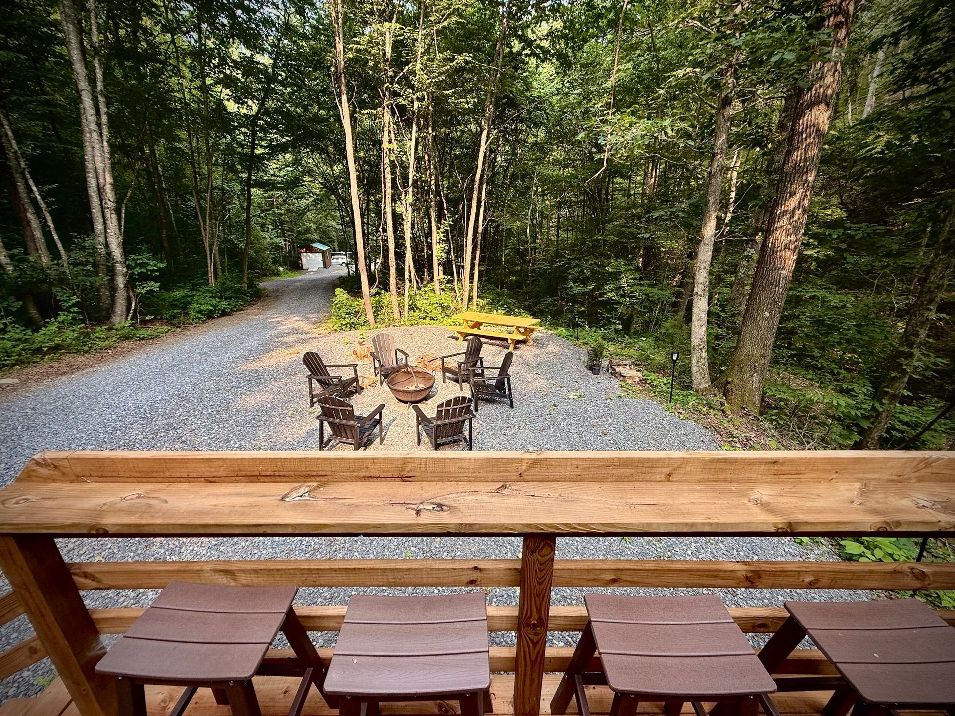 Wooden picnic table with stools beside a forest gravel path, with a fire pit and chairs ahead