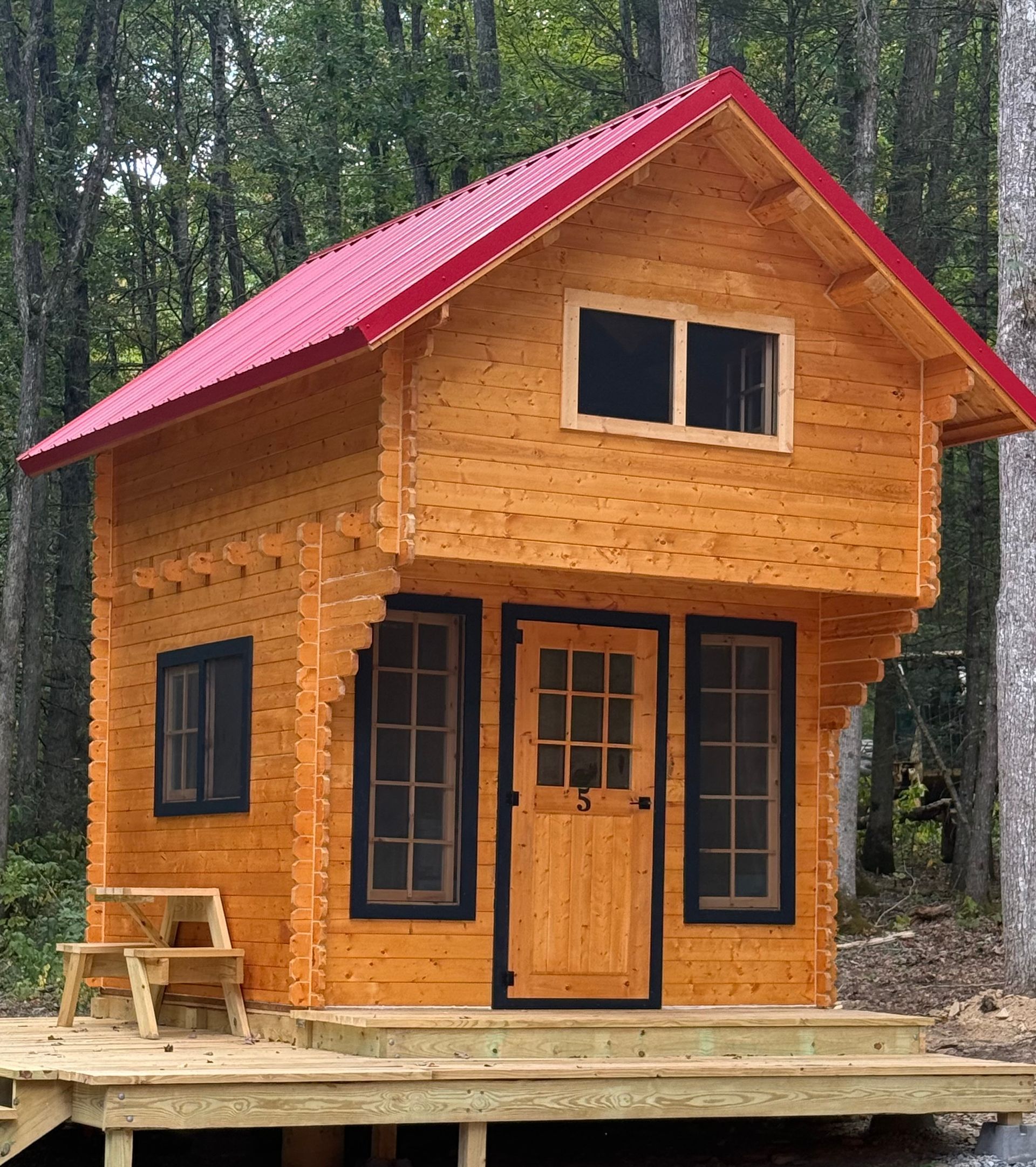 Small wooden cabin with a red roof on a forested site, with a porch and bench.